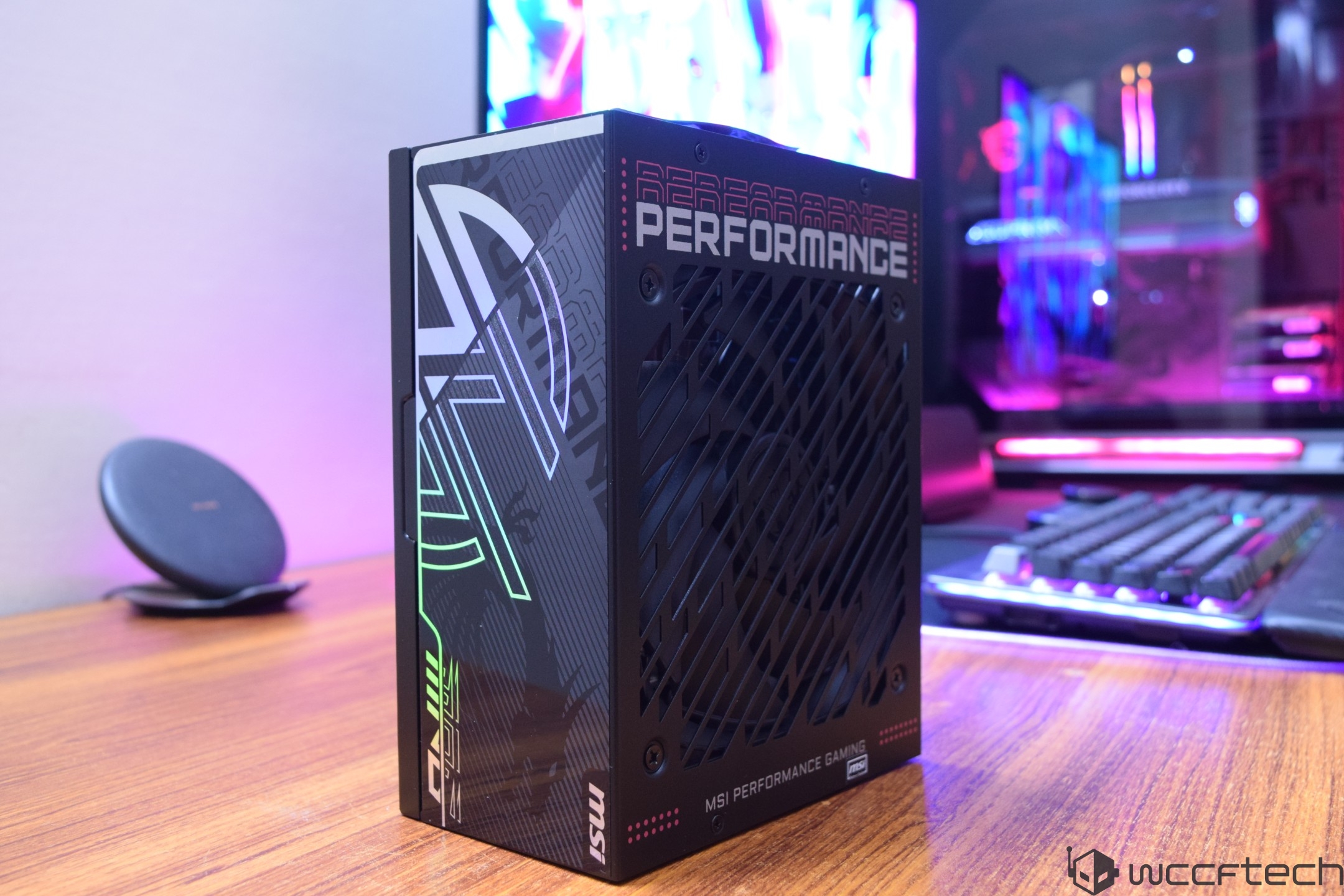 The image shows an MSI Performance Gaming power supply unit featuring the text 'PERFORMANCE' on its side, placed on a wooden desk with a colorful gaming setup in the background.