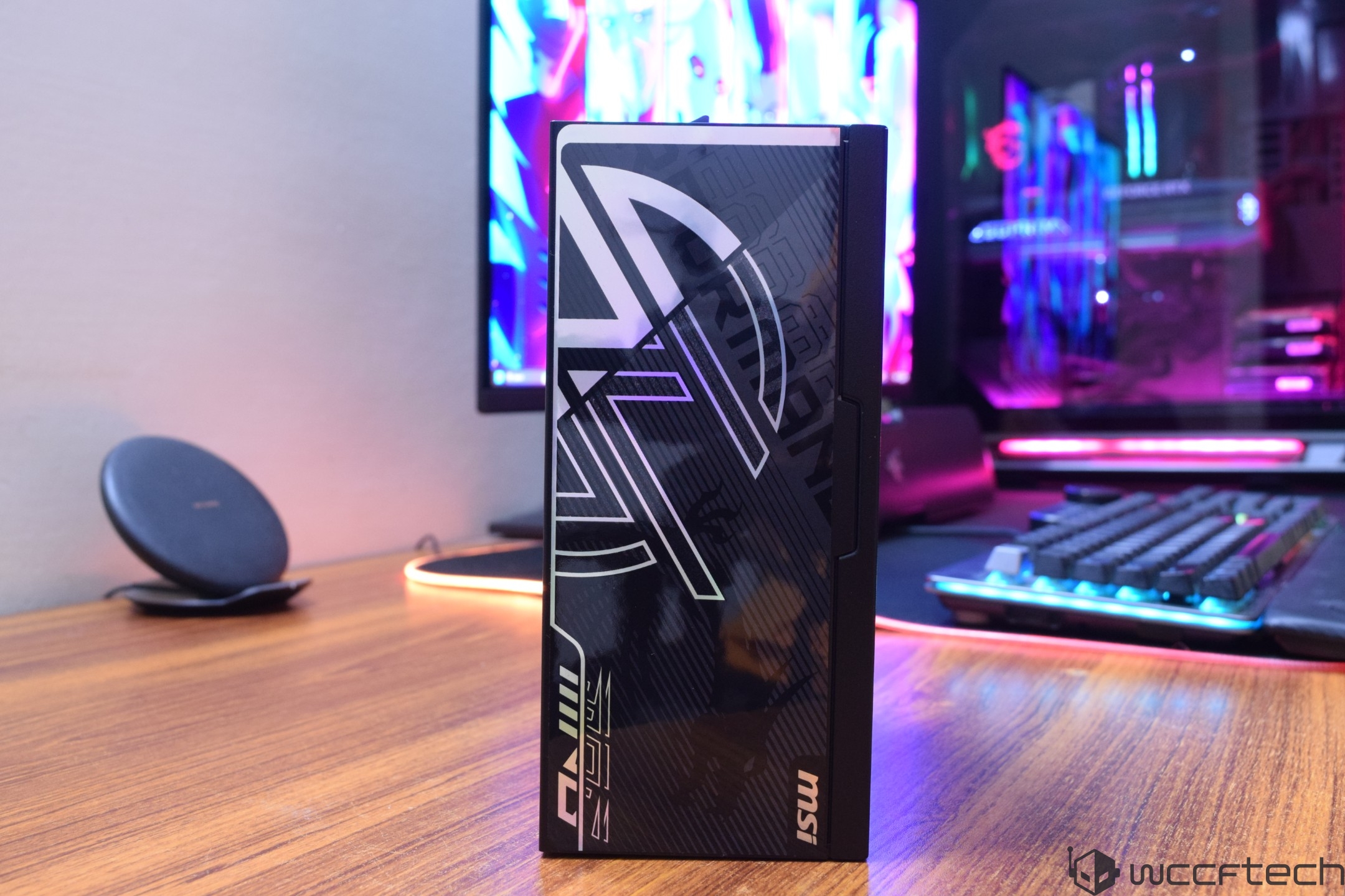 An MSI graphics card box with distinctive graphics is placed on a wooden desk beside a gaming monitor setup.