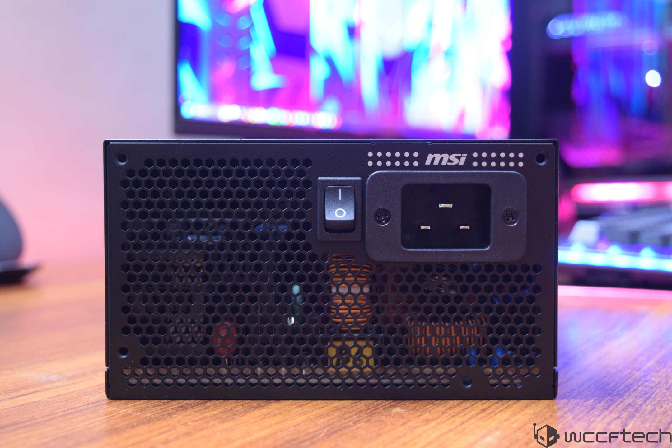 MSI power supply with a honeycomb ventilation pattern, power switch, and input socket on a wooden surface, with a colorful monitor in the background.