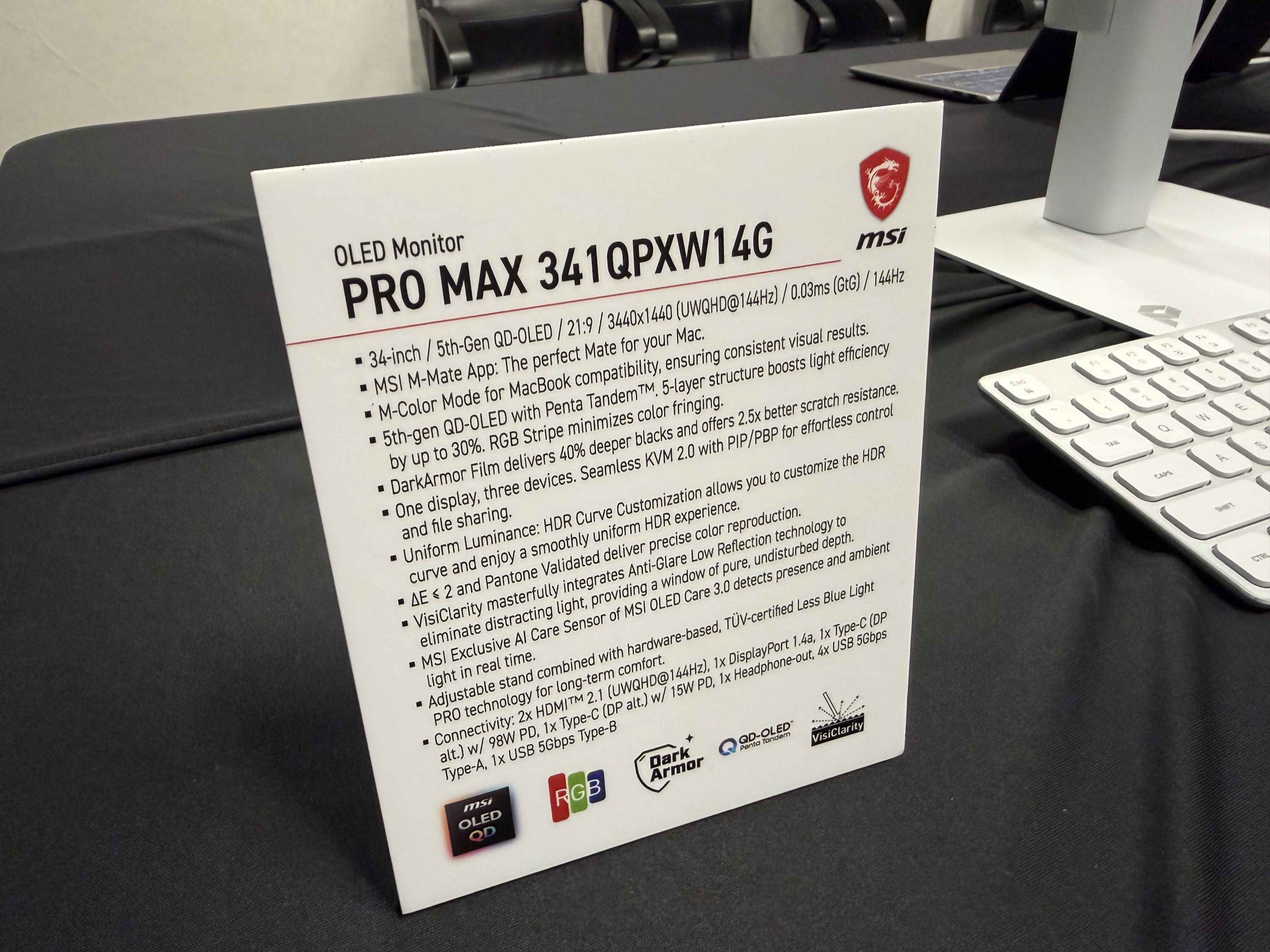 A display card on a table provides specifications for the MSI OLED Monitor PRO MAX 341QPXW14G, highlighting features like a 34-inch 5th-Gen QD-OLED panel, 21:9 aspect ratio, UQWHD resolution at 144Hz, MSI M-Mate App compatibility, and M-Color Mode, with various connectivity options listed.