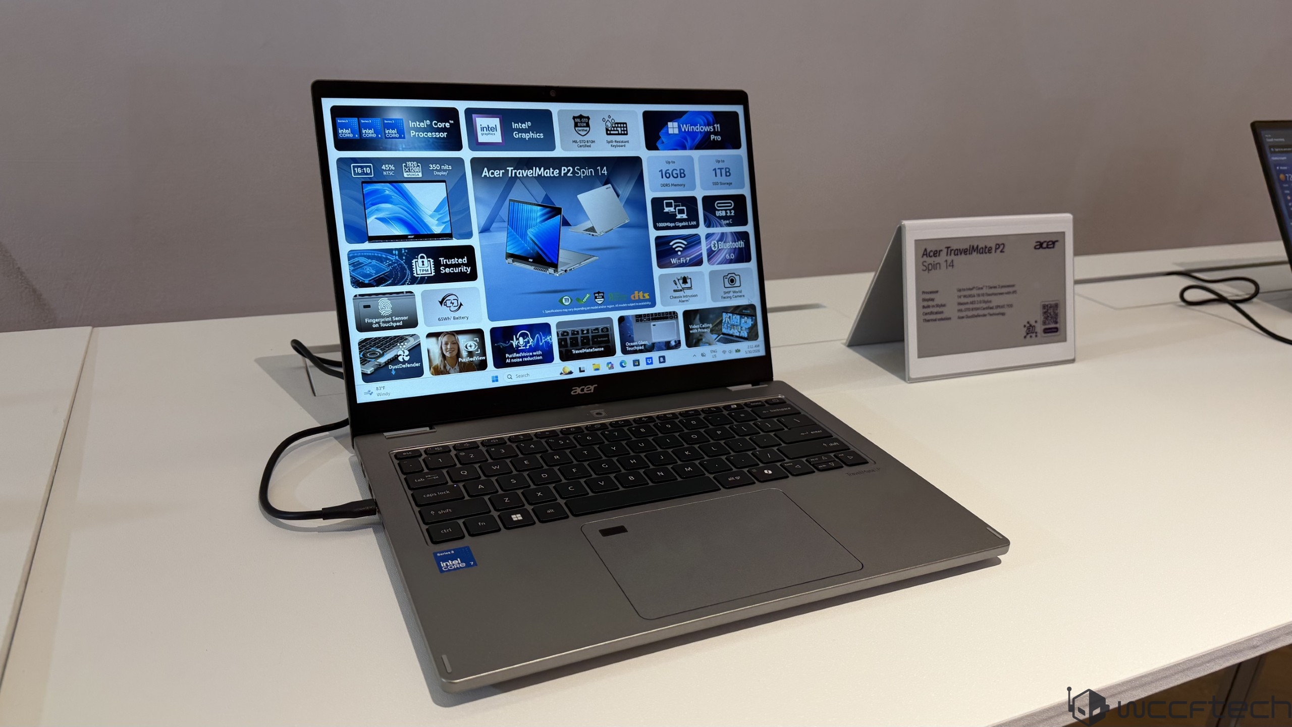 The Acer TravelMate P2 Spin 14 laptop is displayed with a screen showing specifications like '16GB DDR4 Memory' and 'Intel Core Processor,' accompanied by a sign detailing its features.