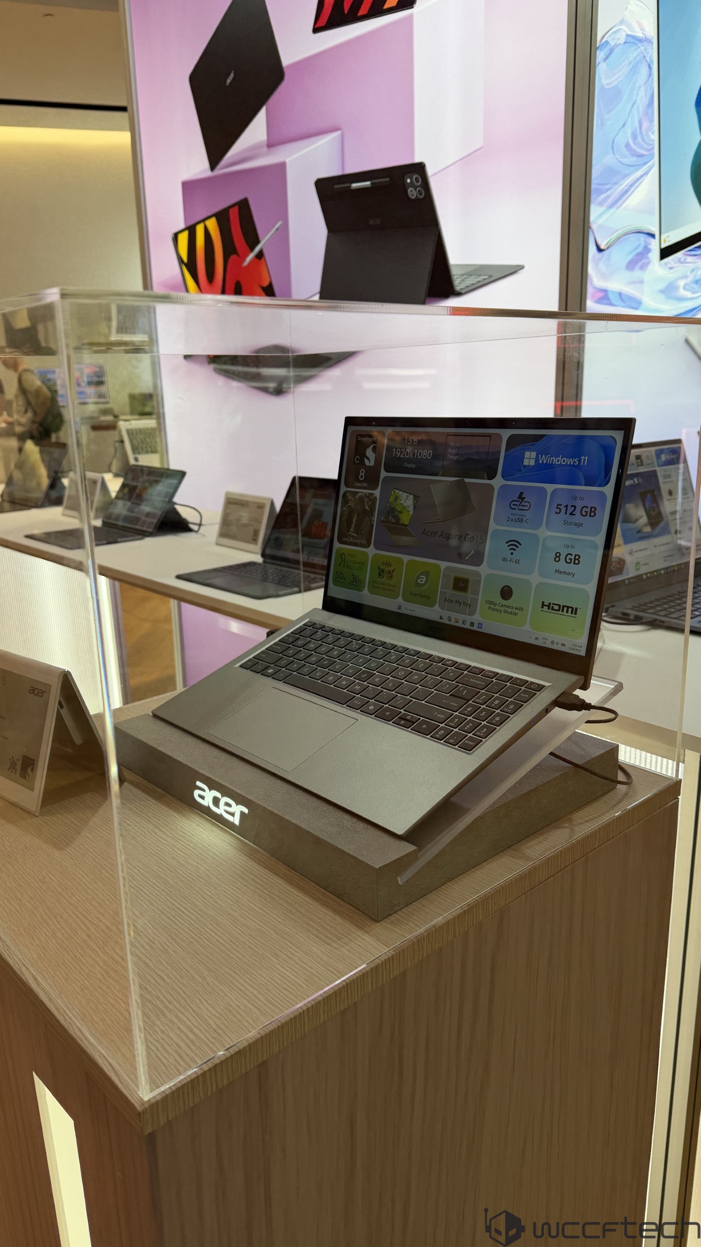 An Acer Aspire Go 15 laptop with an 'Acer Aspire Go 15' label and specifications displayed on-screen, including 'Windows 11,' '512 GB Storage,' '8 GB Memory,' and '1920x1080 Display,' showcased in a glass case.