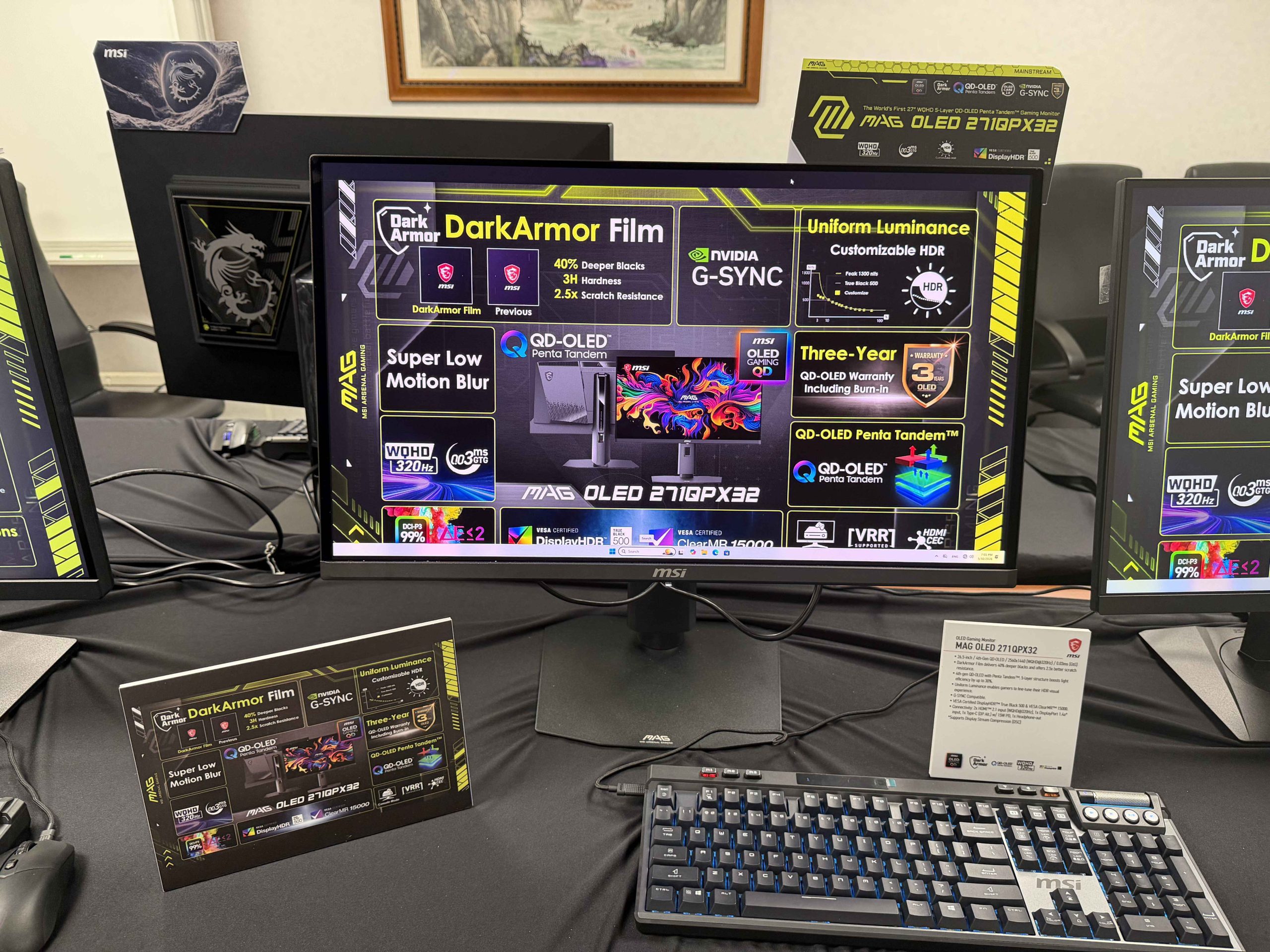 An MSI MAG OLED 271QPX32 monitor displaying features like 'DarkArmor Film,' 'Super Low Motion Blur,' and 'NVIDIA G-SYNC' with a keyboard in front.