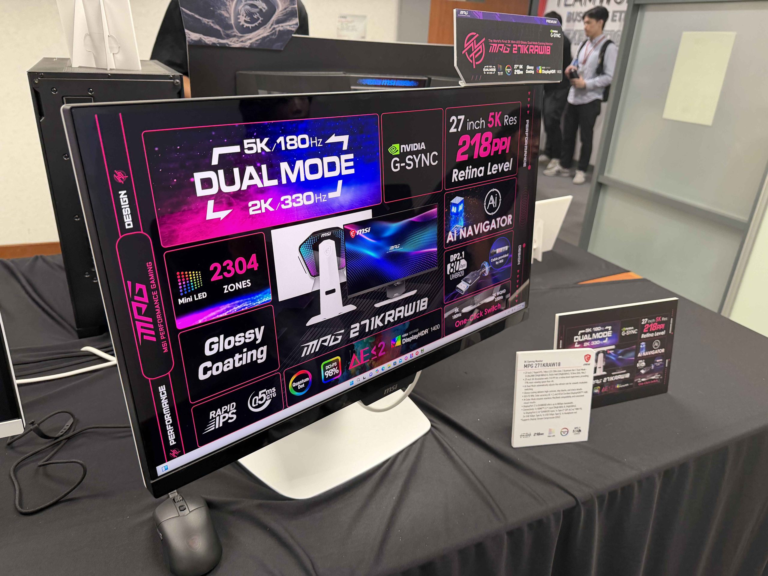A monitor displaying specifications for the MSI MPG 271KRAW18, featuring '5K 180Hz DUAL MODE 2K 330Hz,' 'NVIDIA G-SYNC,' '27 inch 5K Res 218PPI Retina Level,' '2304 ZONES Mini LED,' and 'Glossy Coating.'