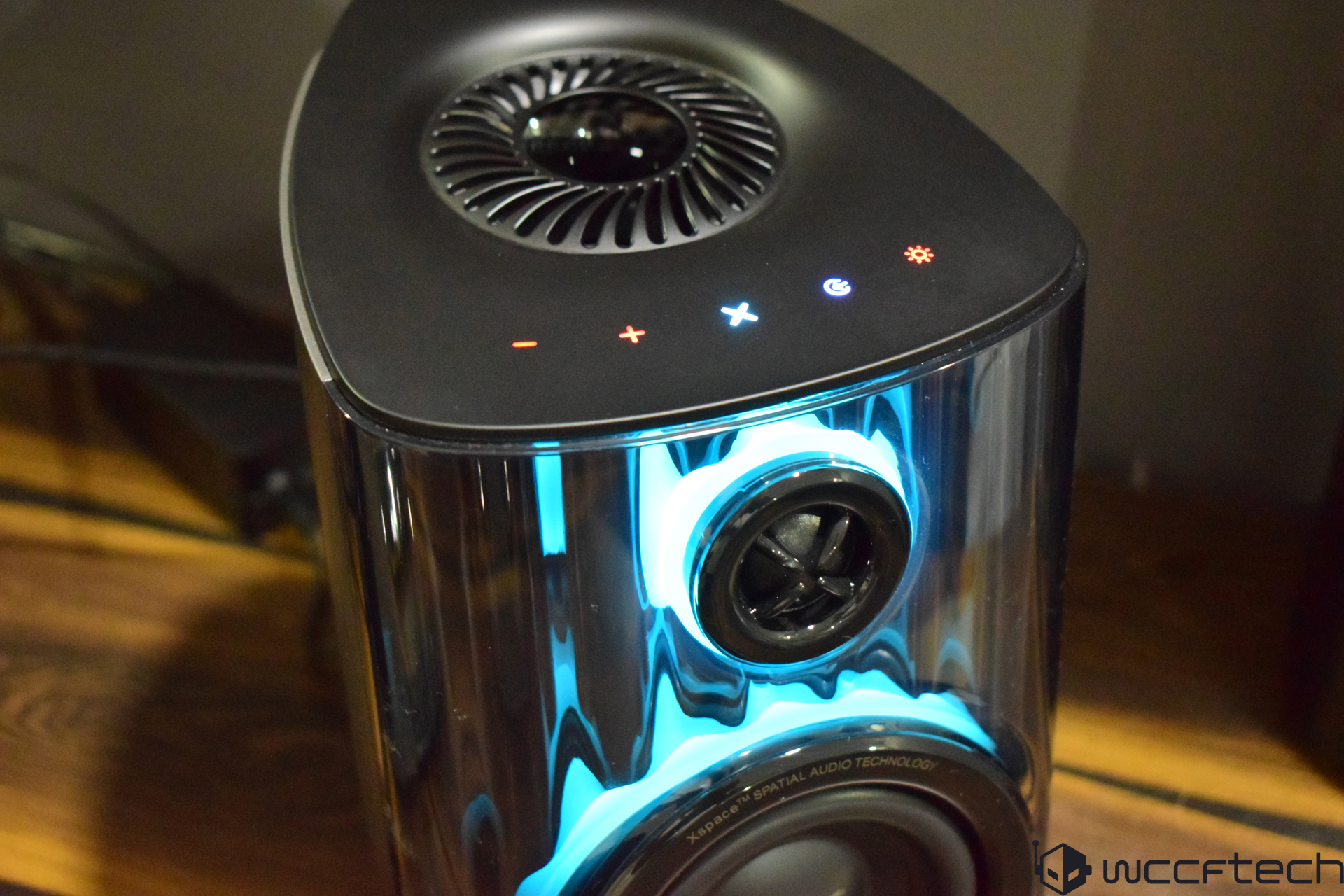 A KEF Mu7 wireless speaker showcasing its glowing blue lighting and top control buttons, with the text 'XSpace Spatial Audio Technology' visible on the front.