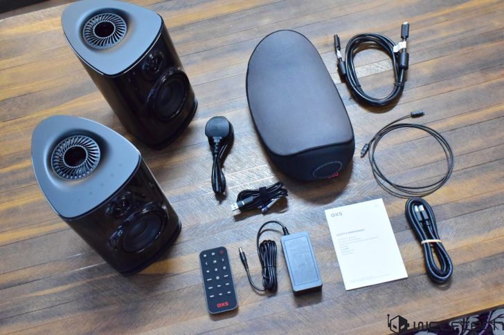 A pair of OXS wireless speakers with accessories, including a remote control, power adapter, and various cables, are displayed on a wooden surface.