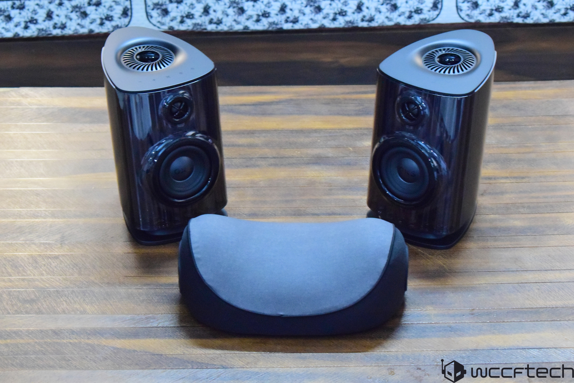 Two KEF wireless speakers are positioned on a wood table with a black fabric-covered accessory between them.