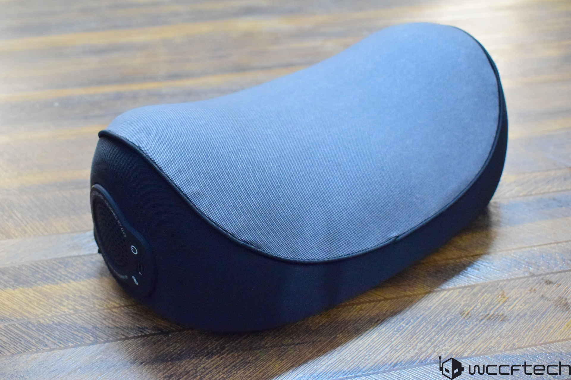 A close-up of a black and gray portable speaker with a fabric cover, showing power and connectivity buttons on the side, placed on a wooden floor.
