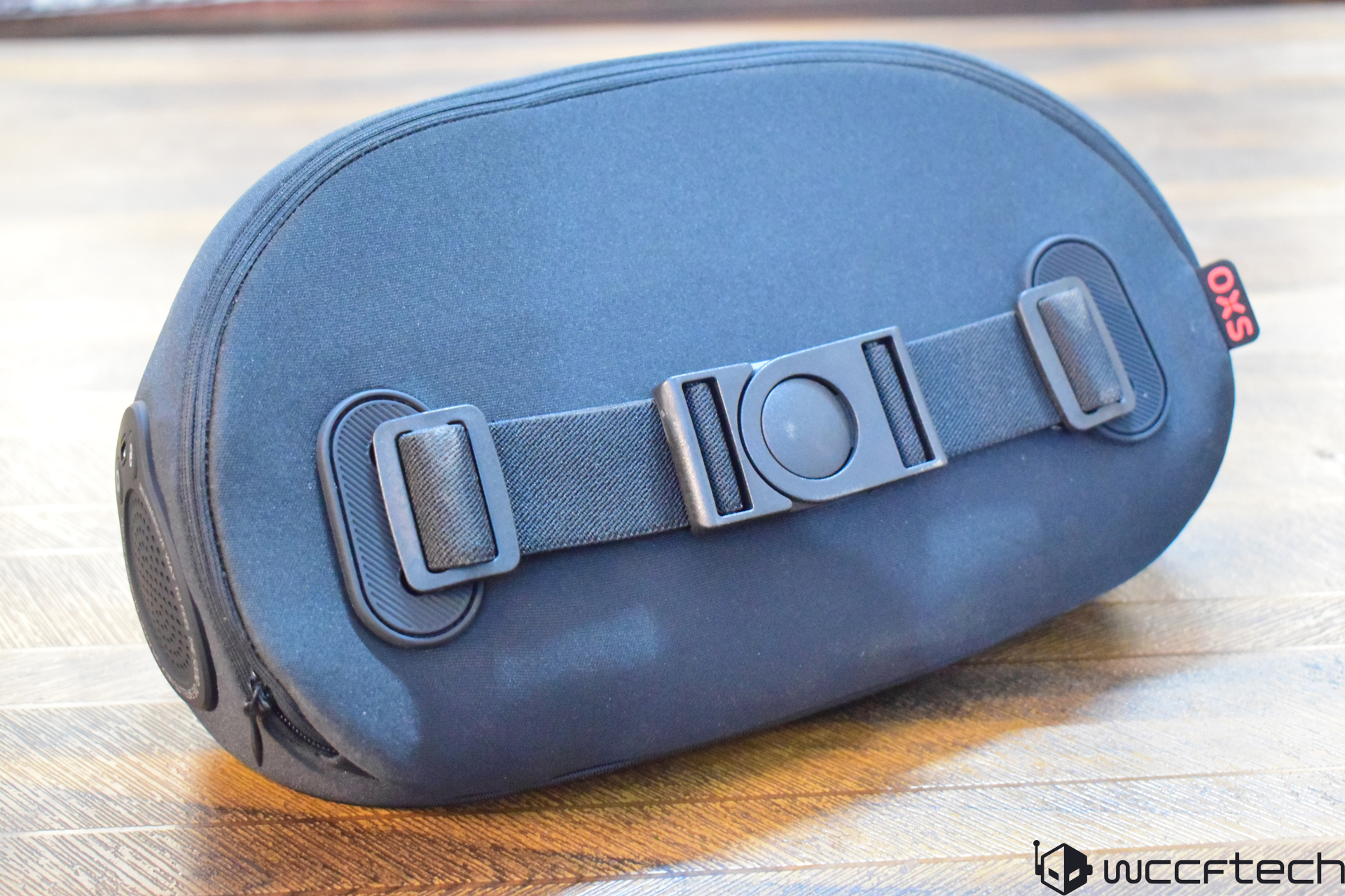 A close-up of a black oval OXS helmet bag with a central buckle and strap on a wooden surface.
