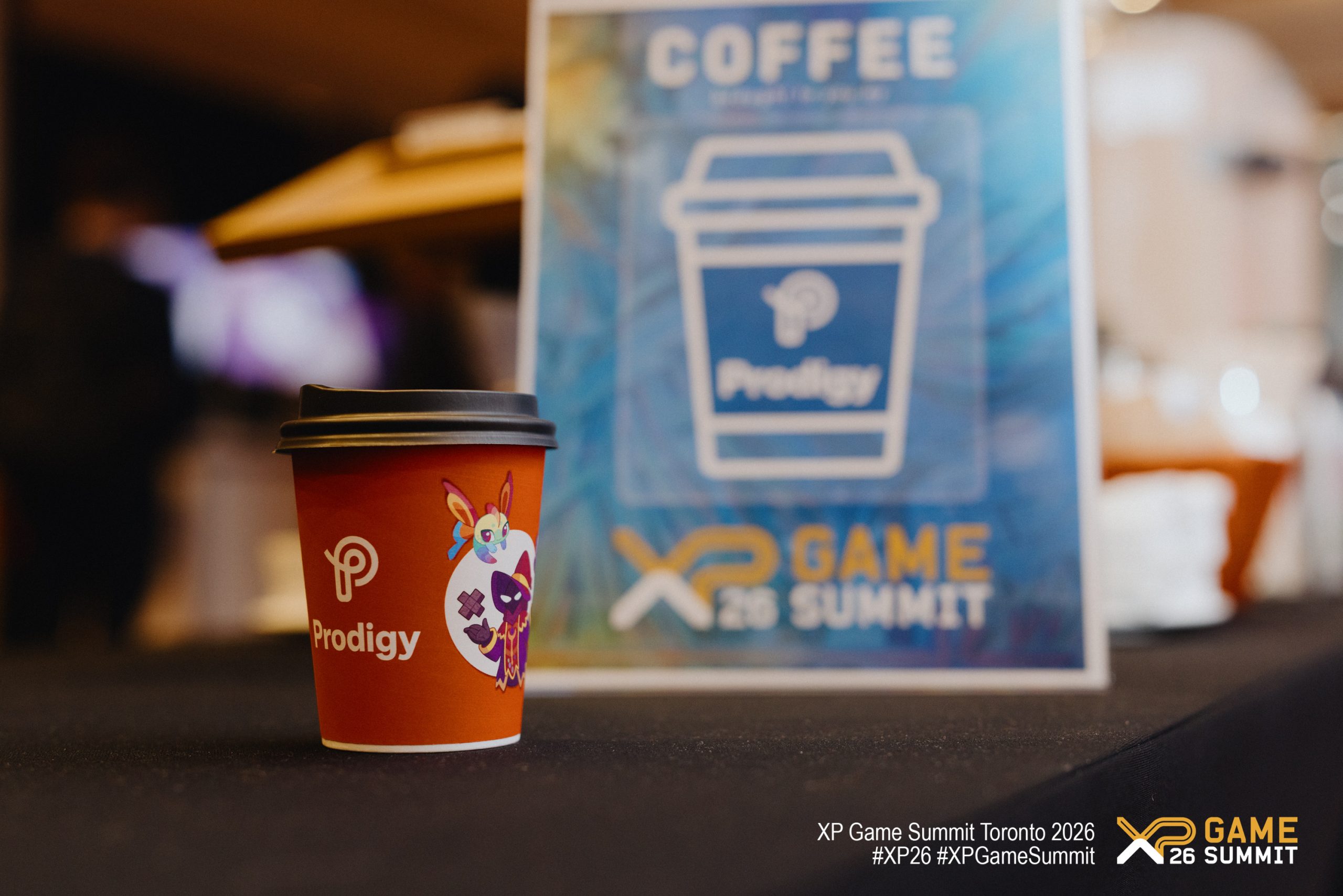 A Prodigy branded coffee cup is placed in front of a sign for the 'XP GAME 26 SUMMIT' at the XP Game Summit Toronto 2026.