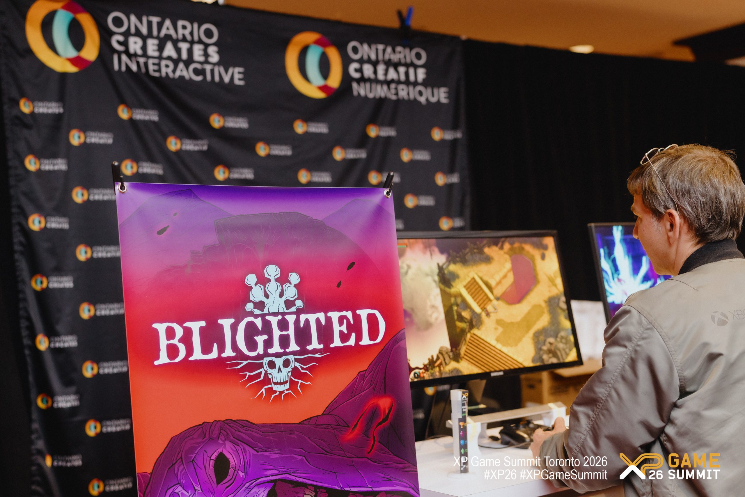 A person is seated at a computer playing a game at the XP Game Summit Toronto 2026, with a prominent poster for the game Blighted and Ontario Creates Interactive signage in the background.