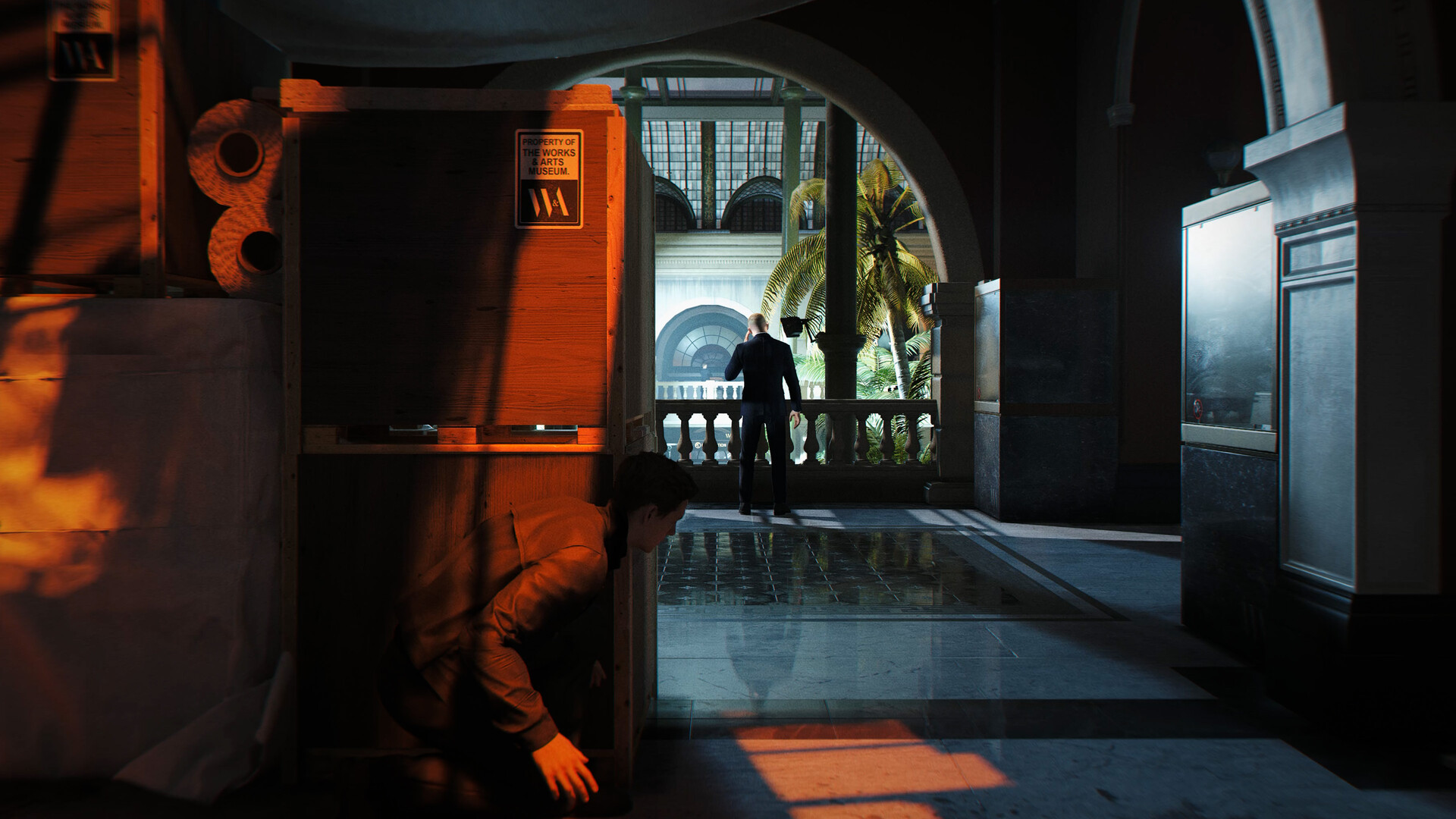 A character in 'Hitman' crouches behind a crate labeled 'PROPERTY OF THE WILKES ART MUSEUM' while another person stands near a balcony.