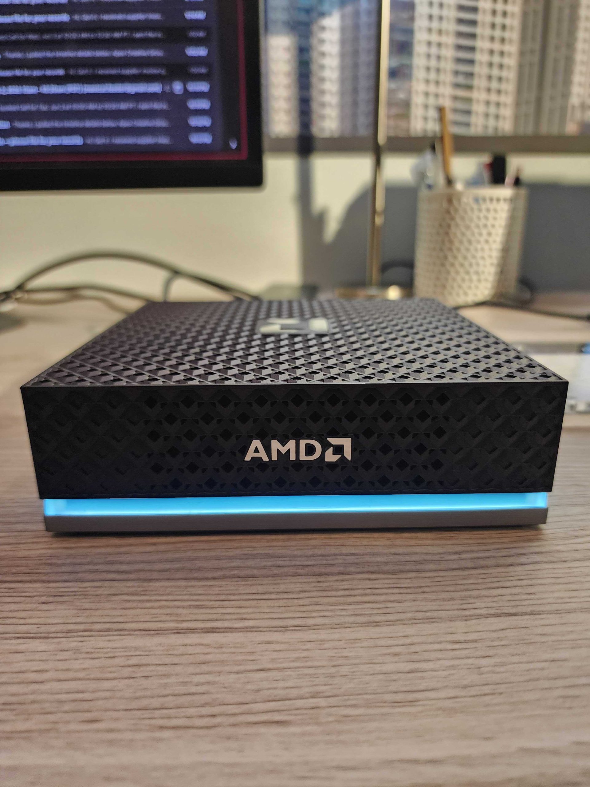 A compact AMD device with a patterned cover and glowing blue base placed on a desk in front of a monitor.
