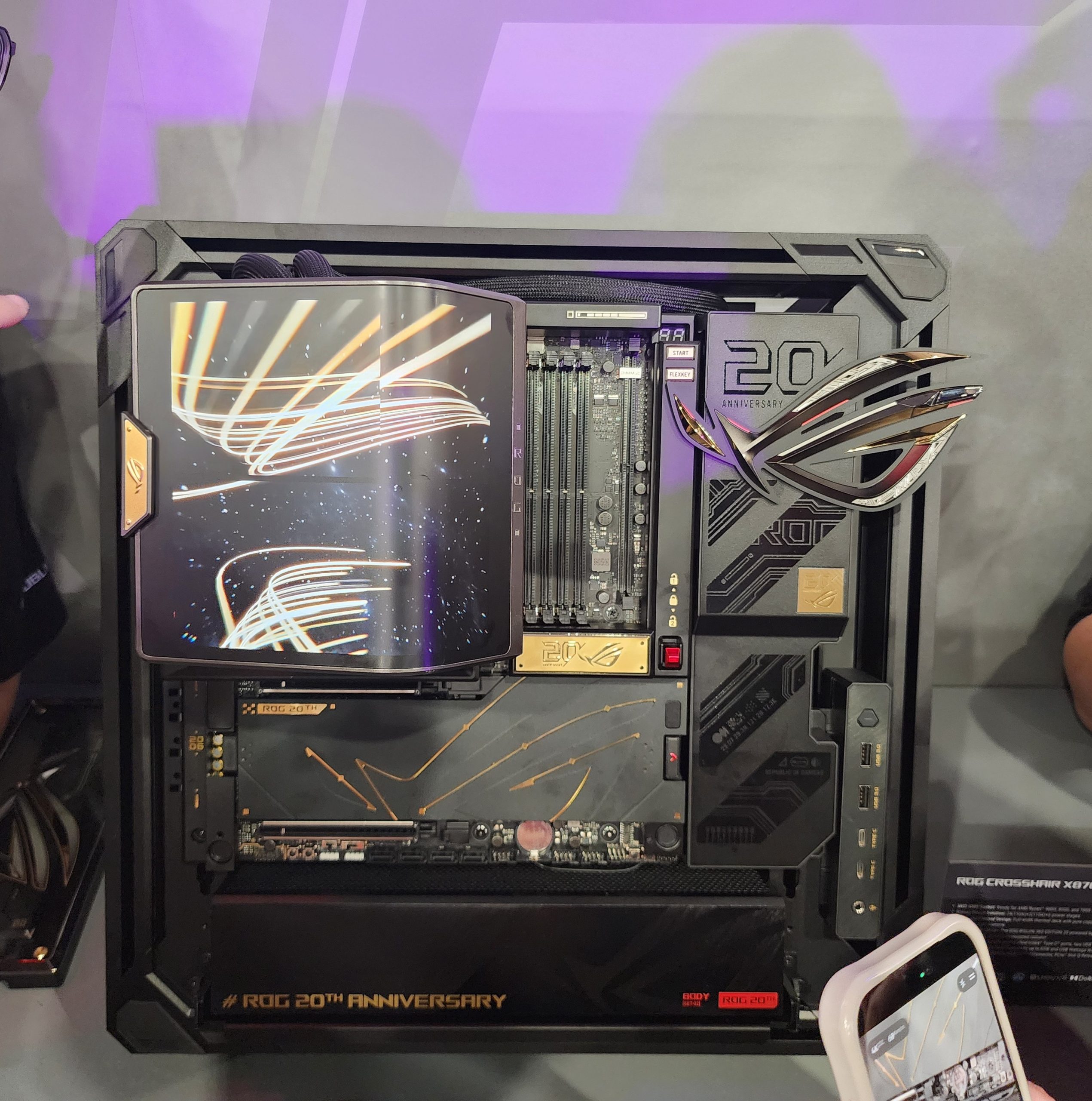 An ASUS ROG Crosshair X670E Gene motherboard is displayed in a custom PC case celebrating 'ROG 20th Anniversary', featuring distinctive gold accents and a large holographic ROG logo.