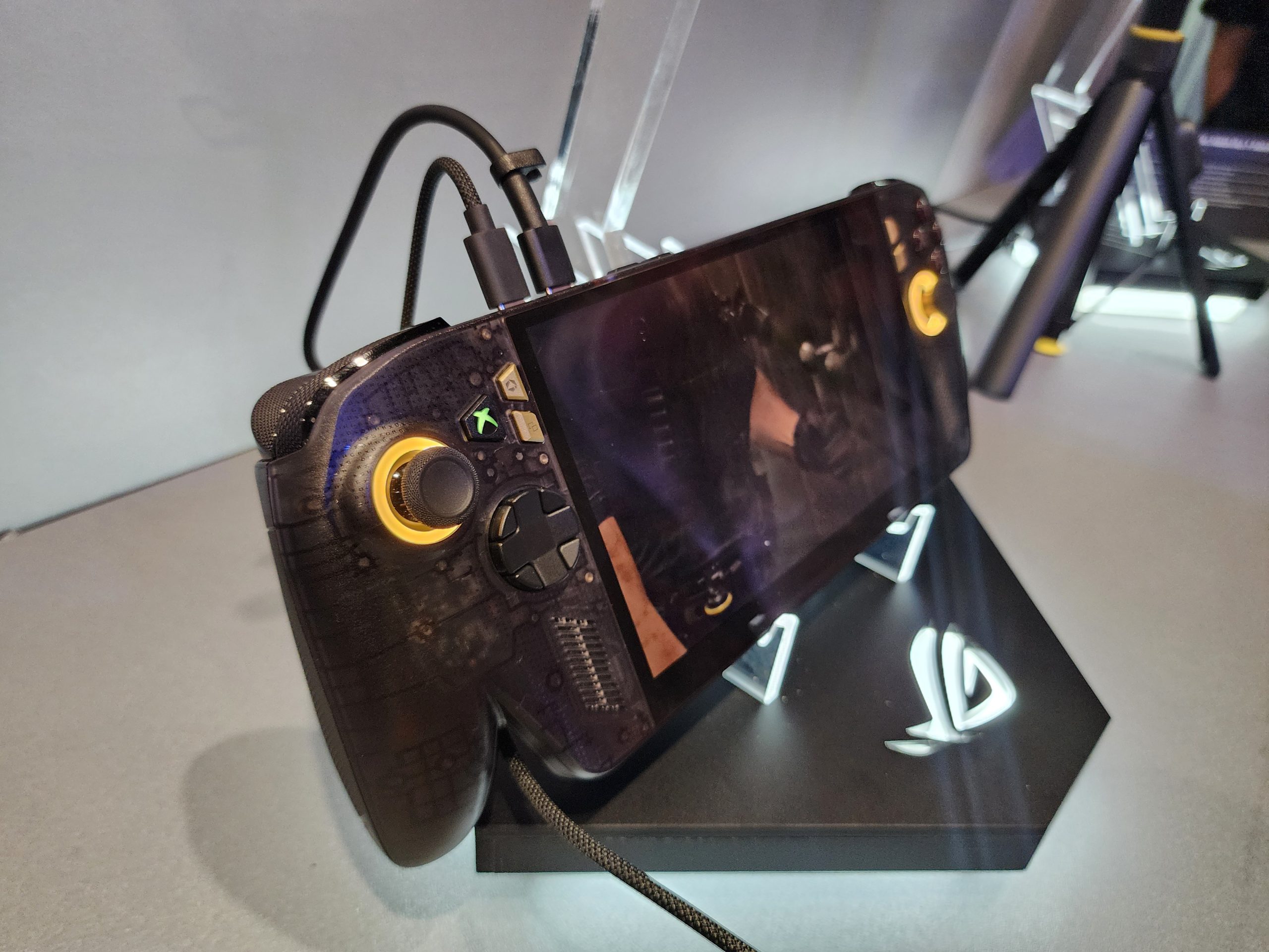 An ASUS ROG Ally gaming handheld device is displayed on a stand with illuminated buttons and a game playing on its screen.
