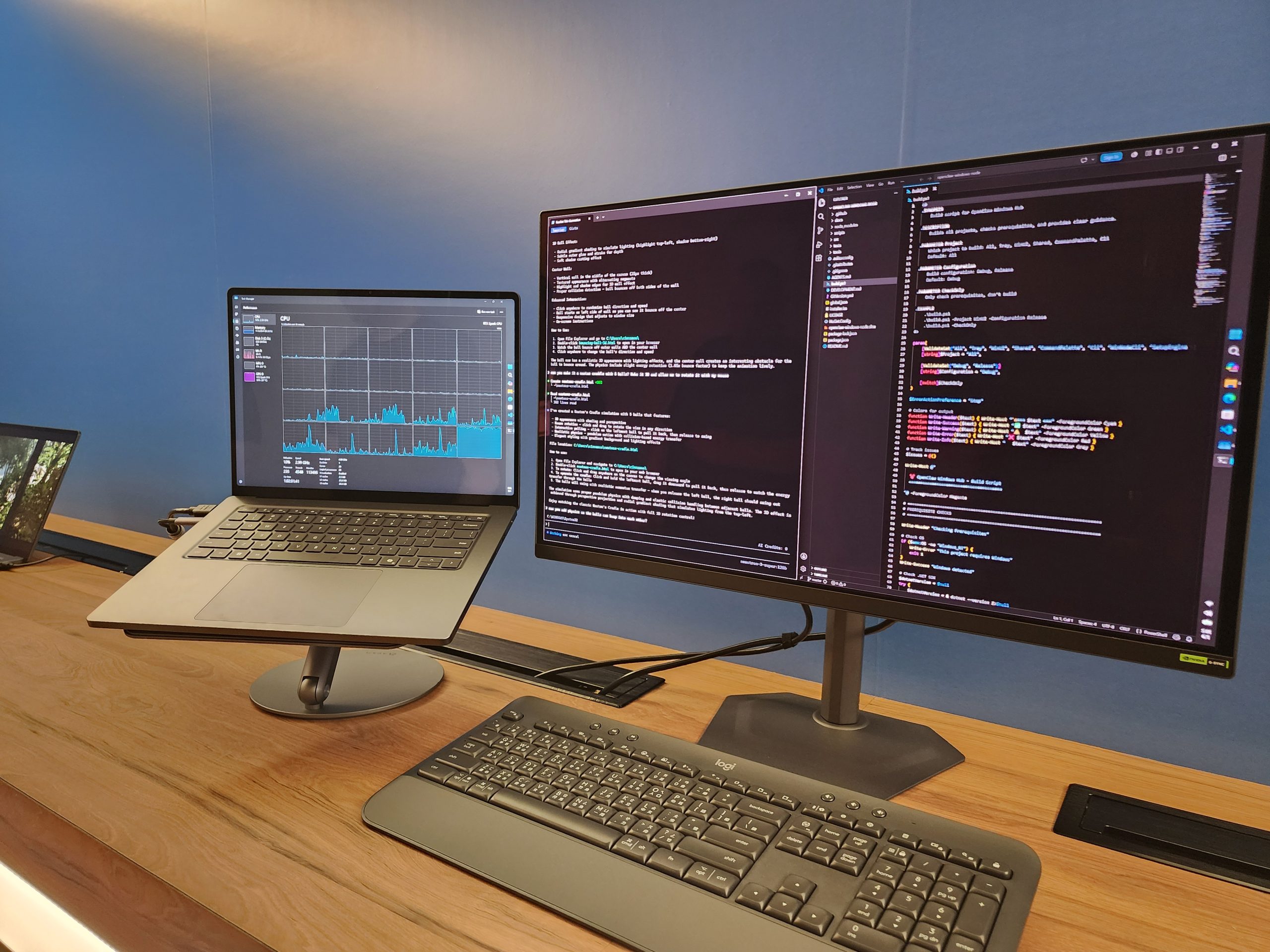 A laptop displaying a CPU performance graph sits next to a monitor showing code in a development environment, both on a wooden desk with a Logitech keyboard.