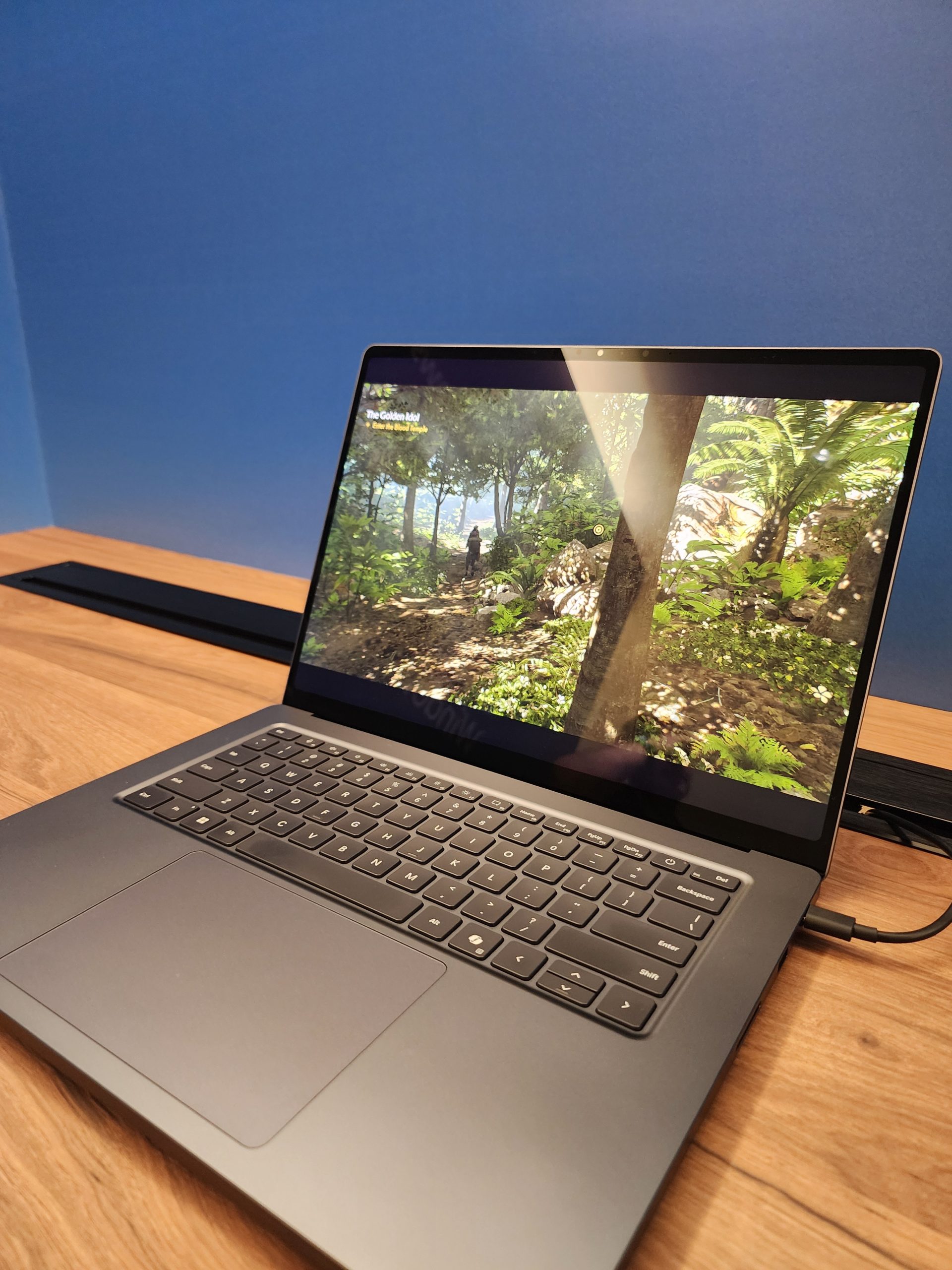 A gaming laptop on a wooden table displays a scene from the game 'The Golden Idol,' showing a lush forest environment on its screen.