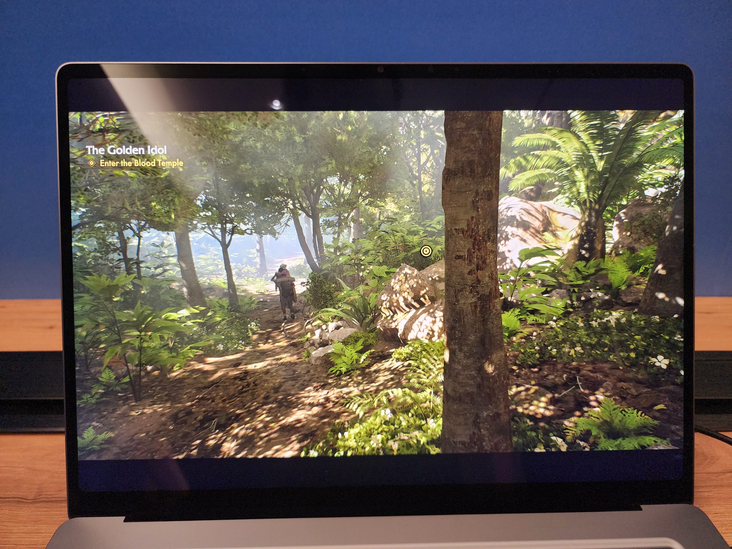A gaming laptop screen shows a character walking through a forest in a game titled 'The Golden Idol,' with the objective 'Enter the Blood Temple.'
