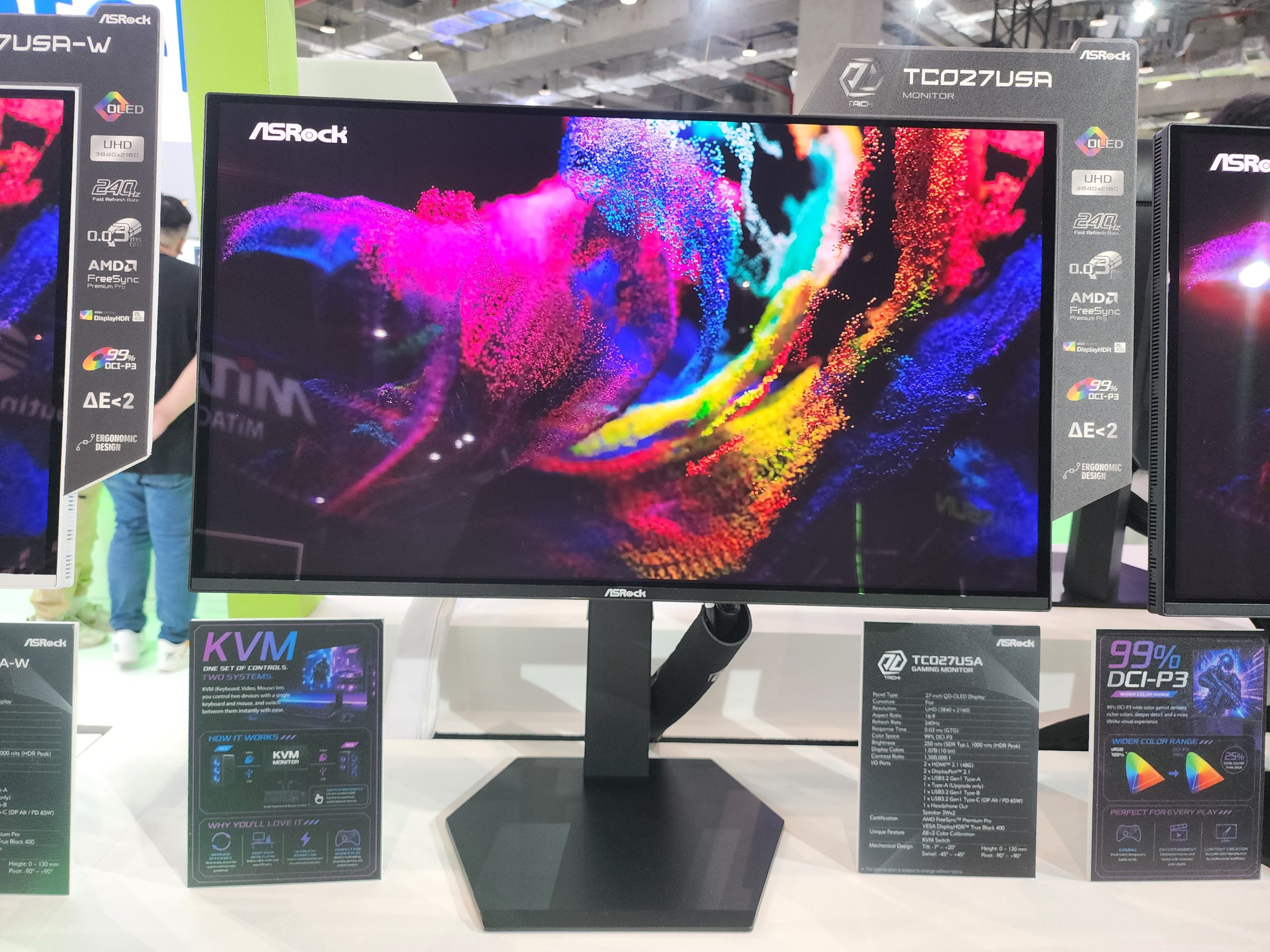 The ASRock TCO27USA QLED monitor is displayed, featuring '99% DCI-P3', '240Hz', and 'AMD FreeSync' capabilities alongside promotional materials highlighting its specifications and KVM switch functionality.