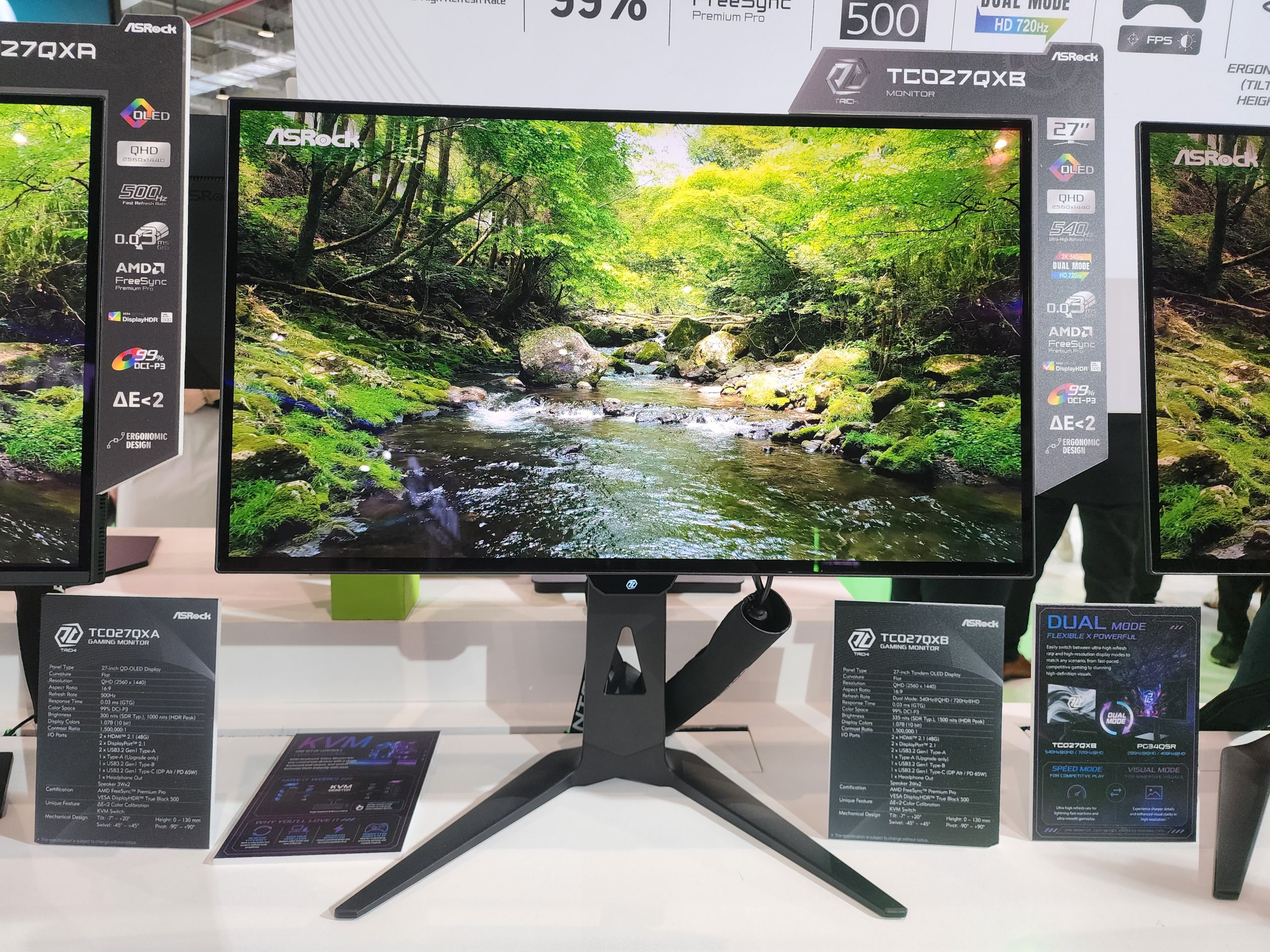 An ASRock TC027QX8 monitor displays a vibrant forest scene and features 'OLED,' 'QHD,' '540,' 'AMD FreeSync,' and '99% DCI-P3' on its side panel.