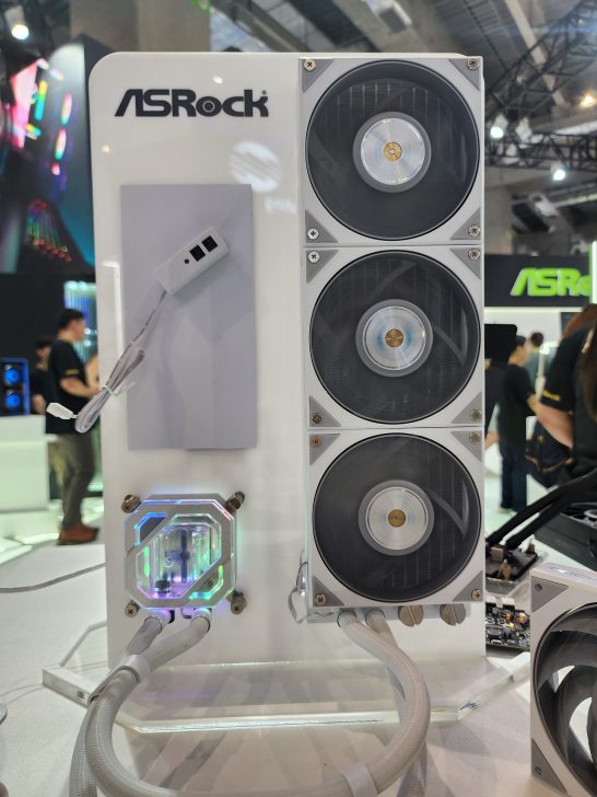 An ASRock cooling system with three large fans and RGB-lit water block on display at a tech event.