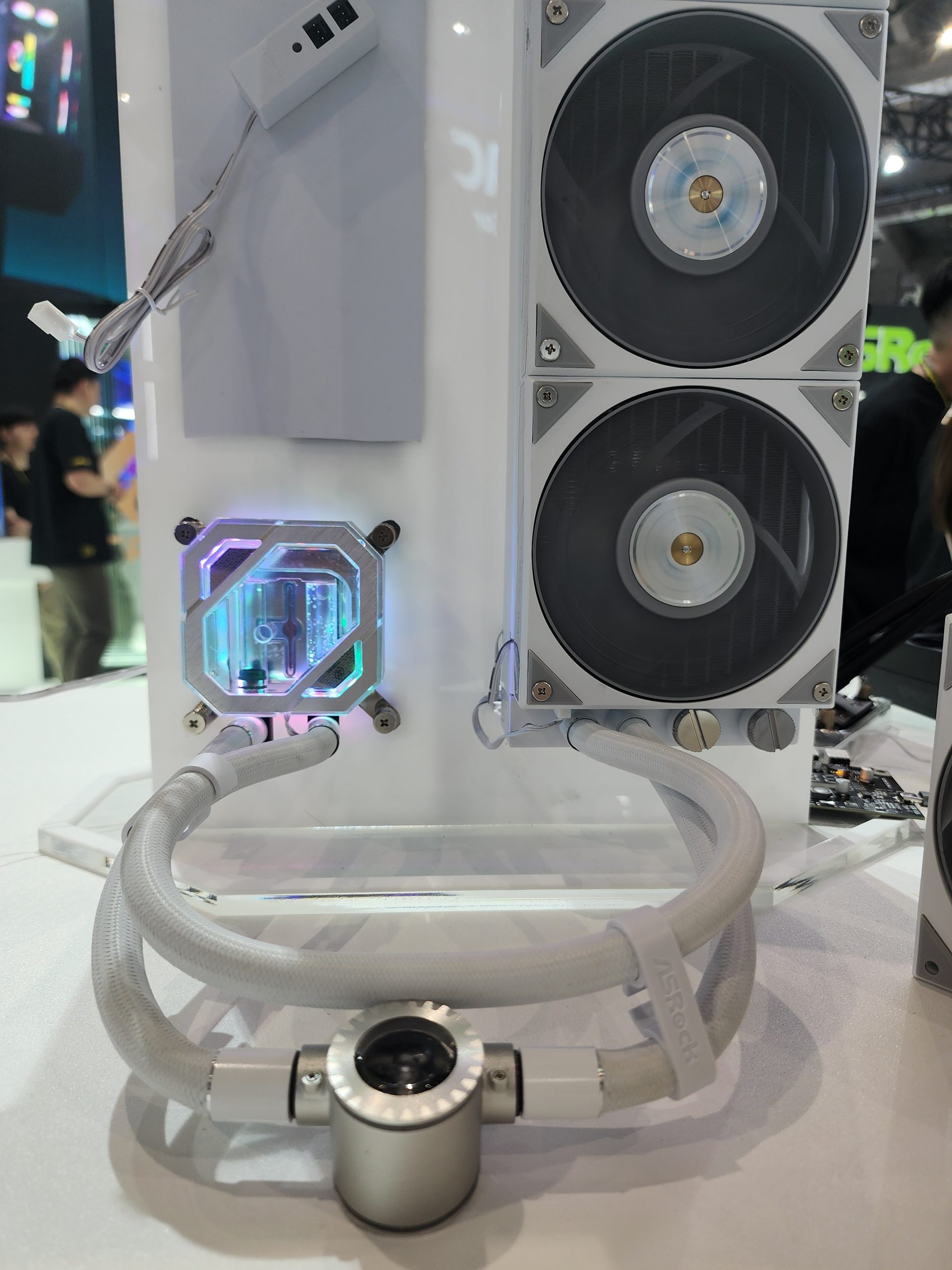 An ASRock liquid cooling system is displayed, featuring two fans, a pump with braided tubing, and illuminated components.