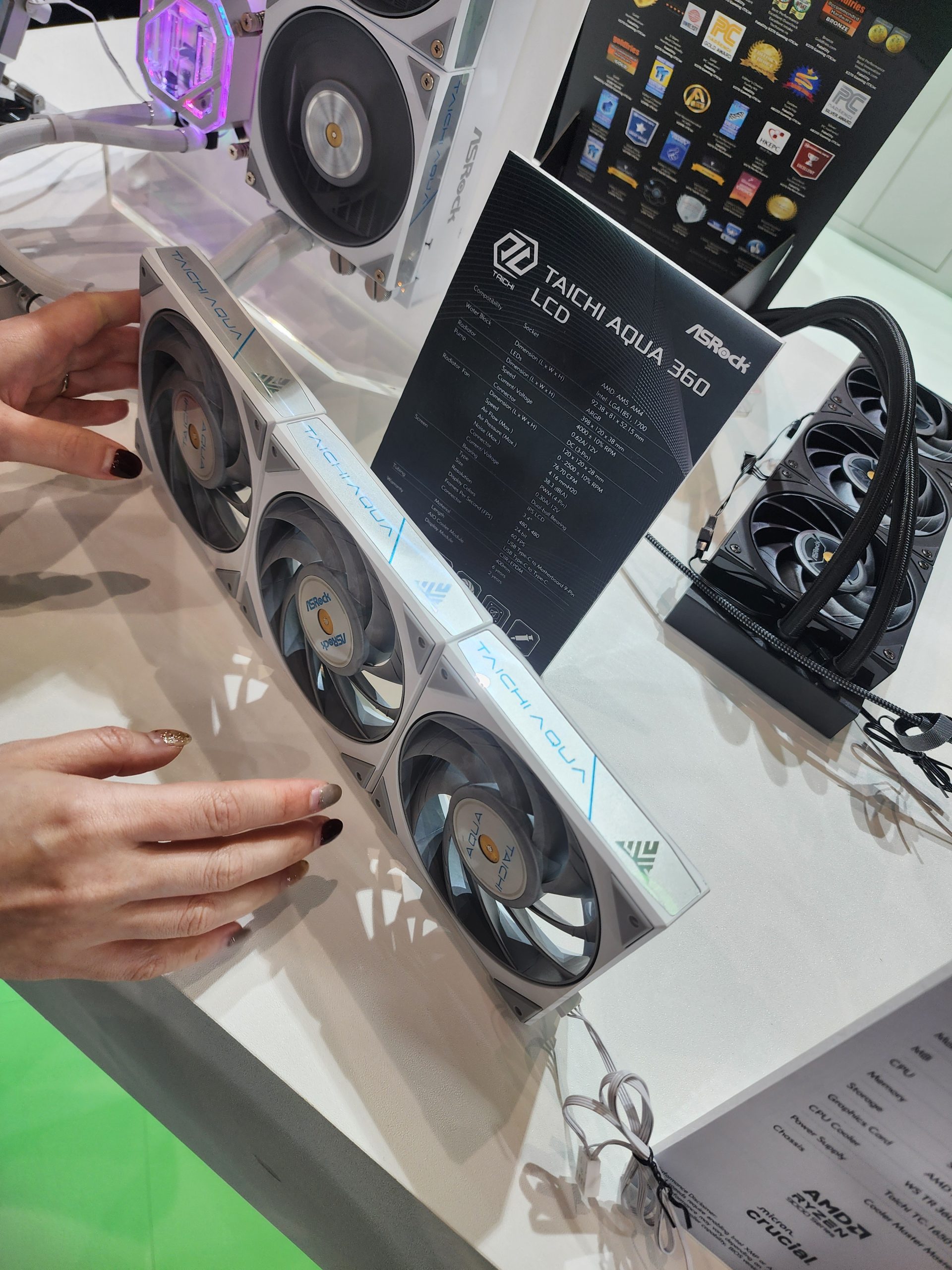 An ASRock Taichi Aqua 360 LCD cooling system is displayed with a specification sheet, while a person adjusts the RGB-lit fan unit.