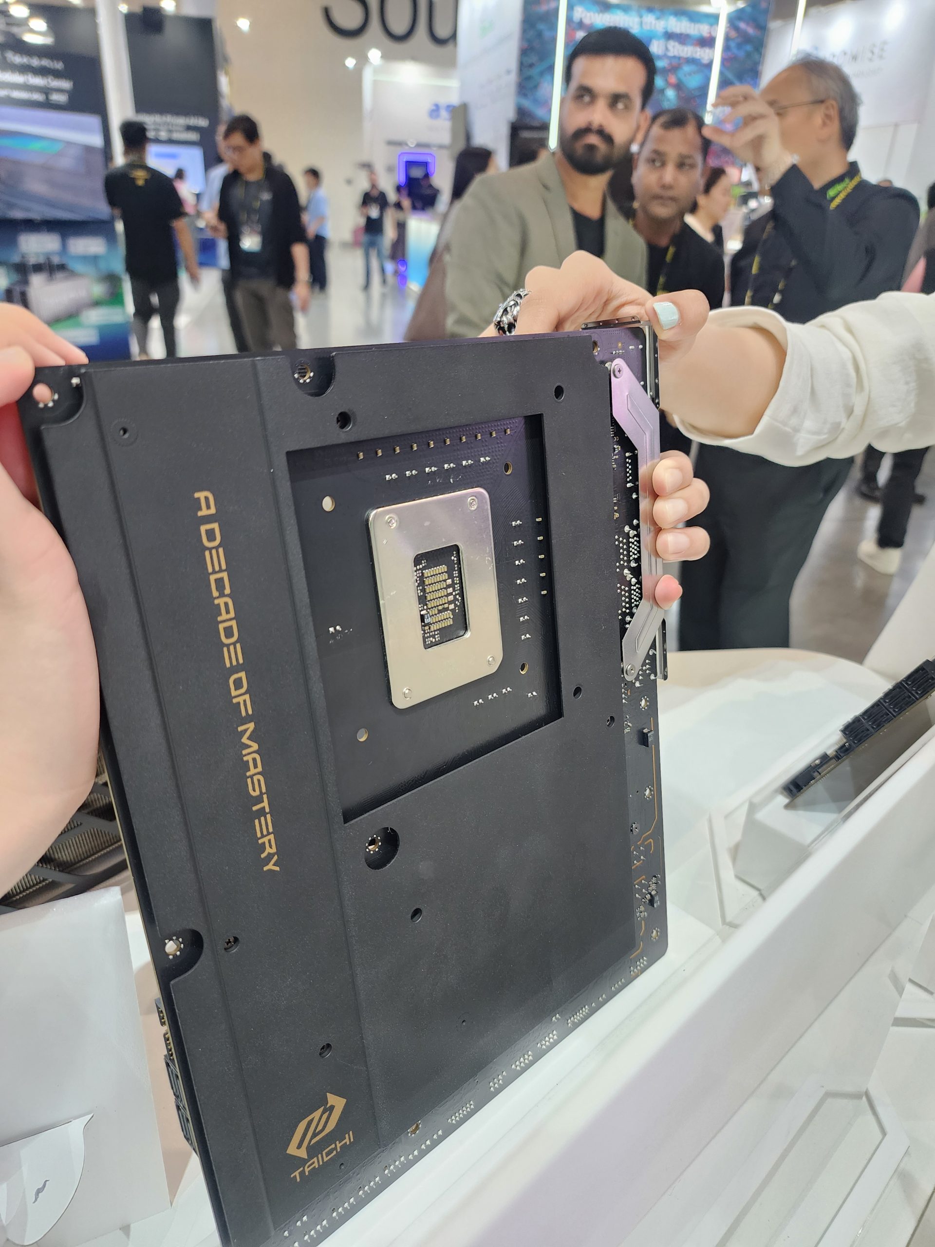 A person holding an ASRock Taichi motherboard with the text 'A DECADE OF MASTERY' visible, in an exhibition setting with attendees in the background.