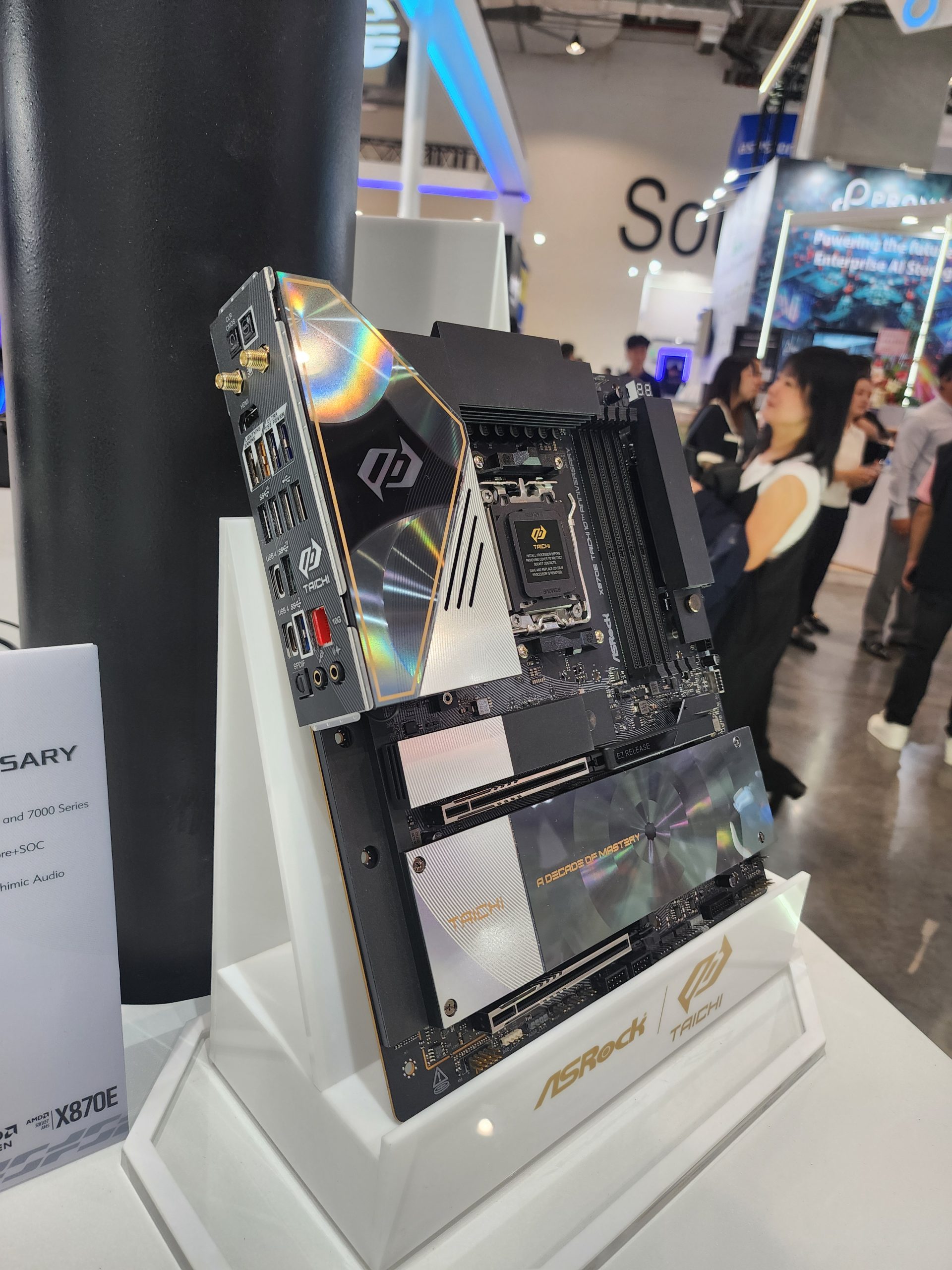 An ASRock Taichi X670E motherboard is displayed at an event, featuring 'A DECOROUS MASTERY' on its design.