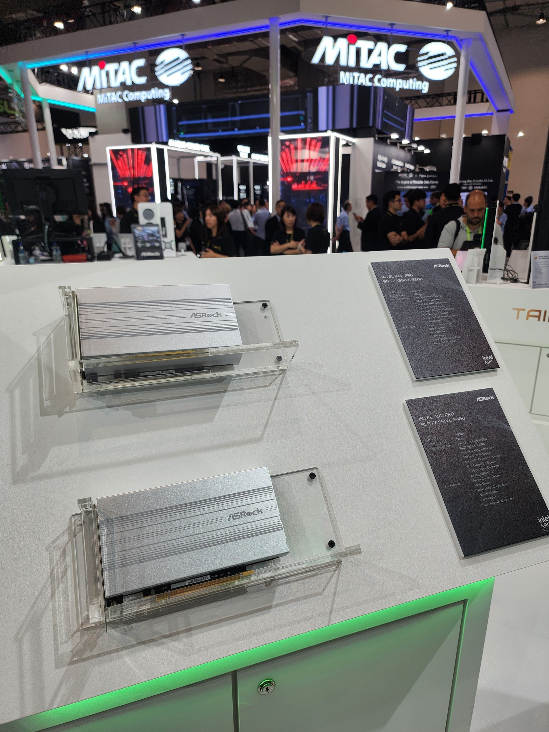 Two ASRock Intel ARC Pro graphics cards displayed on a counter at a tech event, with a visible ASRock information card detailing specifications, and the MiTAC Computing booth in the background.