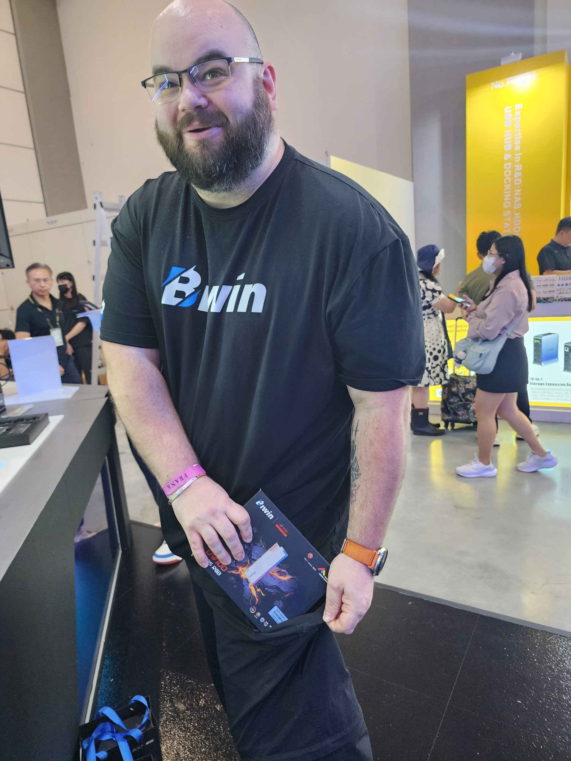 A person wearing a 'Powerwin' t-shirt holds a product box labeled 'Powerwin' at a tech event, with a yellow display in the background mentioning 'USB HUB DOCKING STATION'.