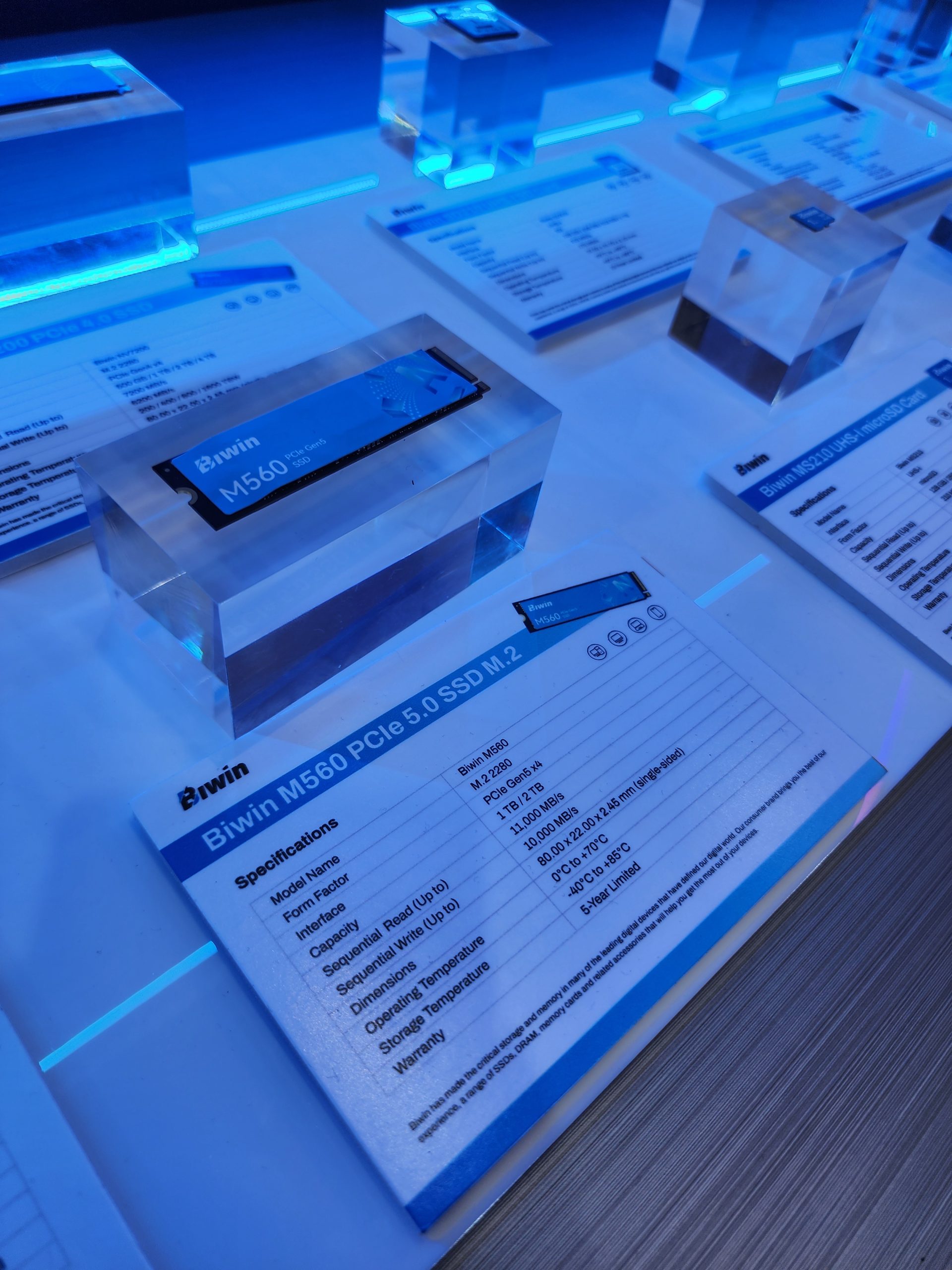 A display showcasing the Biwin M560 PCIe 5.0 SSD M.2 alongside a detailed specifications card stating features like PCIe Gen 5, storage capacity of 1/2 TB, and sequential read and write speeds of up to 12,000 MB/s and 10,000 MB/s respectively.