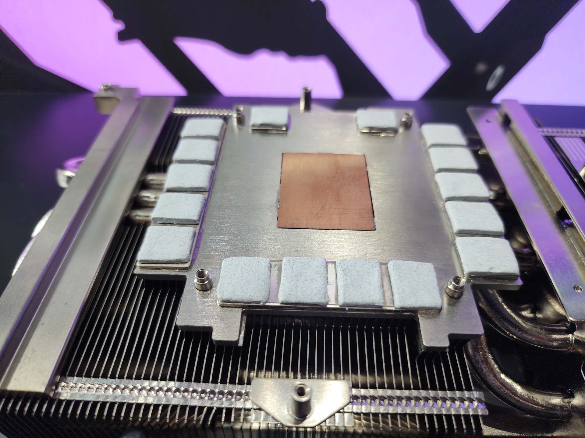 A close-up of a GPU cooling system with a copper heat spreader and thermal pads on metal fins, set against a purple background.