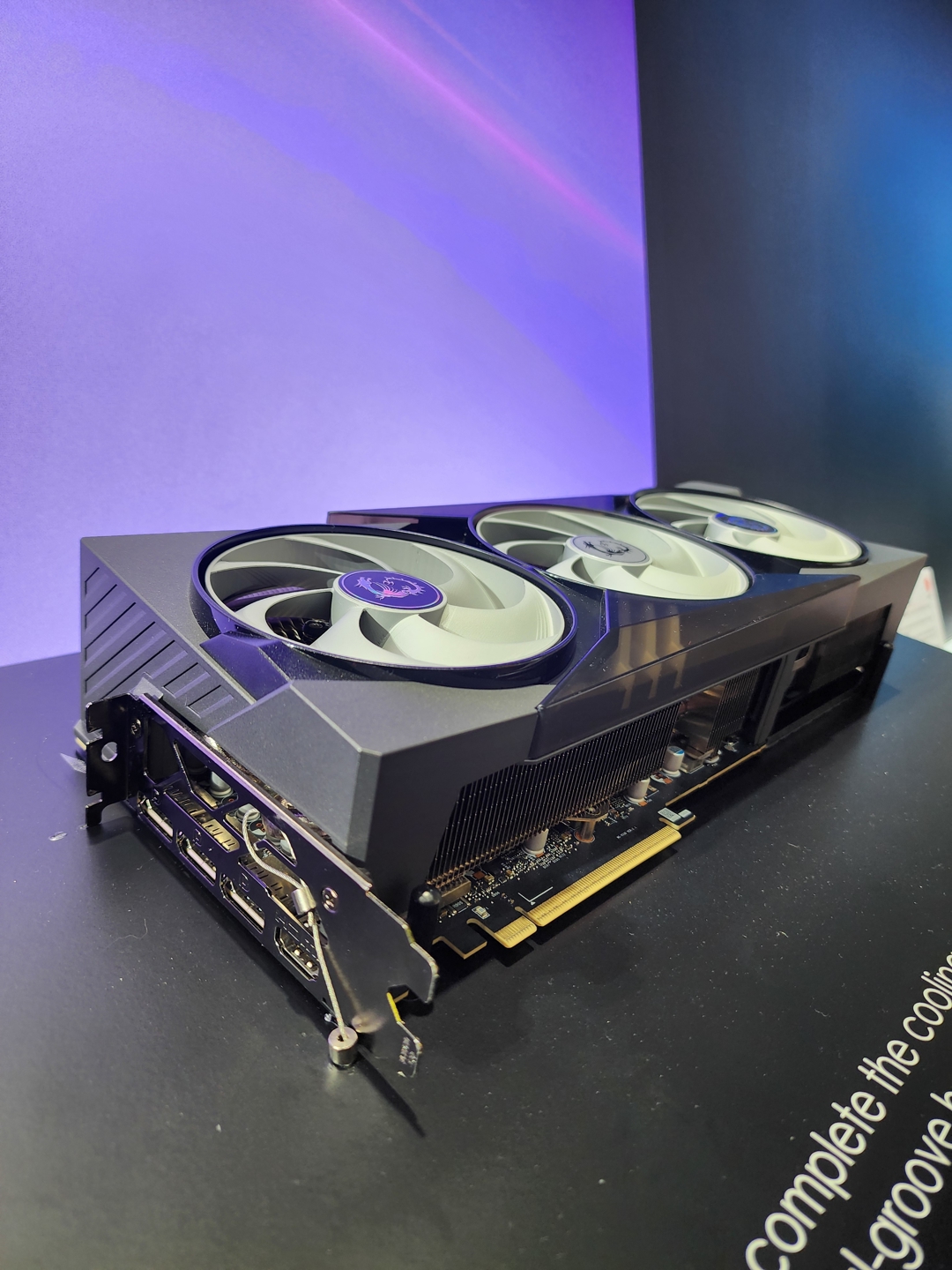 A black GPU with three fans on the top, each featuring a logo in the center, is displayed against a purple gradient background.