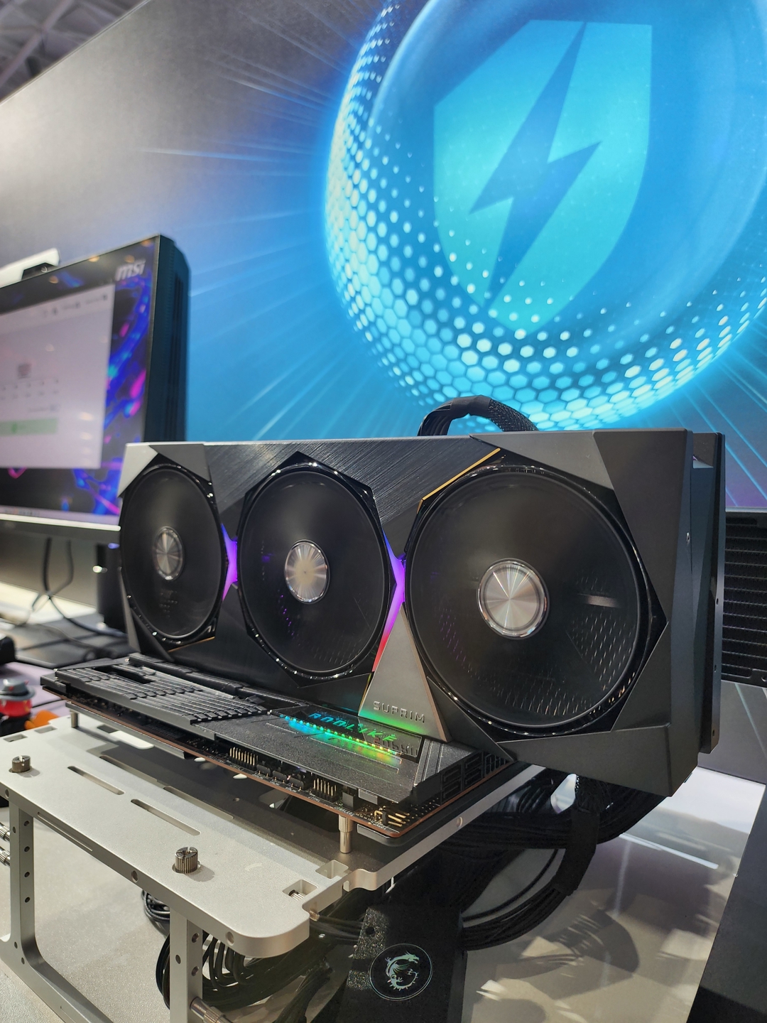 A large MSI graphics card with three fans and 'SUPRIM' branding installed in a test bench setup, with an MSI monitor visible in the background.