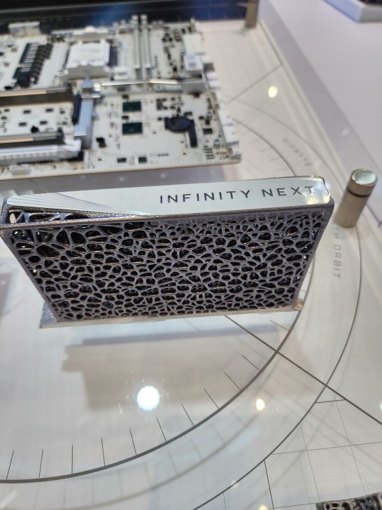 A hardware component branded as 'INFINITY NEXT' is displayed on a stand, with a motherboard visible in the background.
