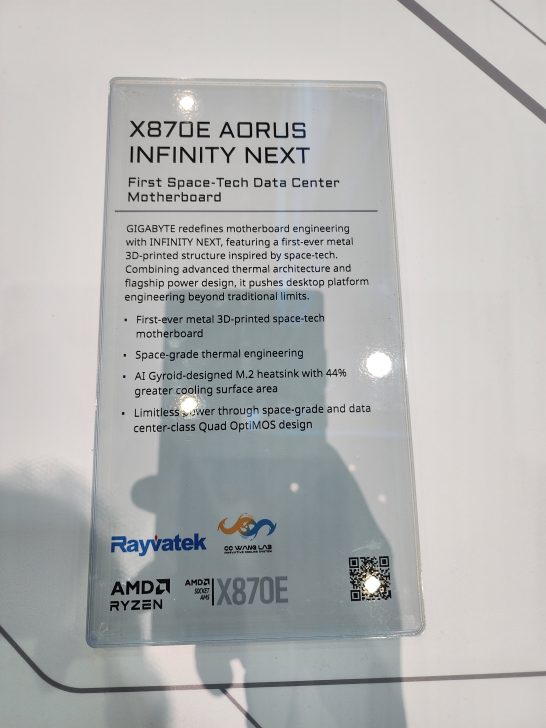 A plaque details the Gigabyte X870E Aorus Infinity Next as the first space-tech data center motherboard, featuring a metal 3D-printed structure, AI gyroid-designed M.2 heatsink, and space-grade thermal engineering, with logos from Rayvatek, OC Wana Lab, and AMD Ryzen.