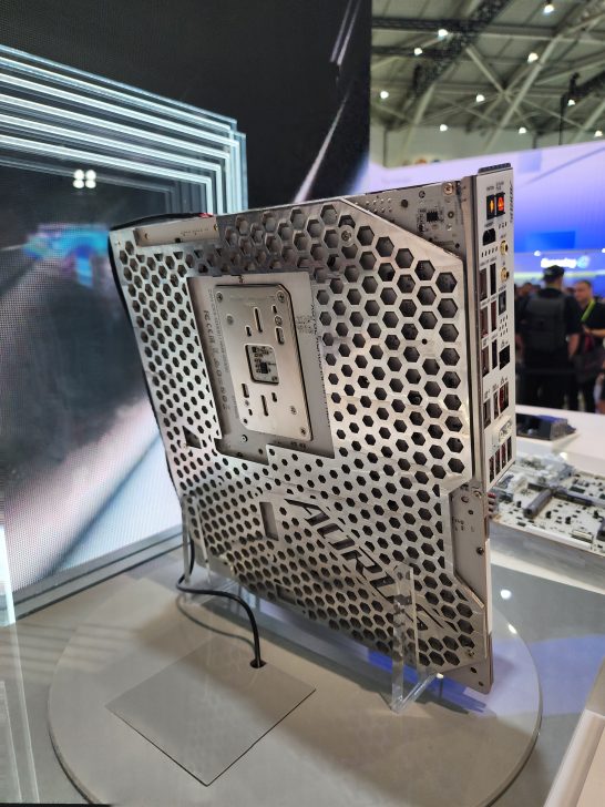 The image shows an ASUS motherboard with a hexagonal mesh design displayed vertically at a tech exhibition.