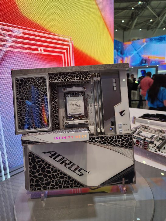 A Gigabyte AORUS motherboard labeled 'INFINITY NEXT' is displayed on a table at an exhibition, featuring prominently its unique design and branding.