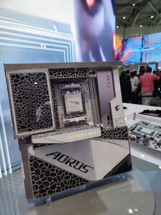 A Gigabyte Aorus Infinity Next motherboard is displayed at a tech event with intricate lattice cooling and branding visible.