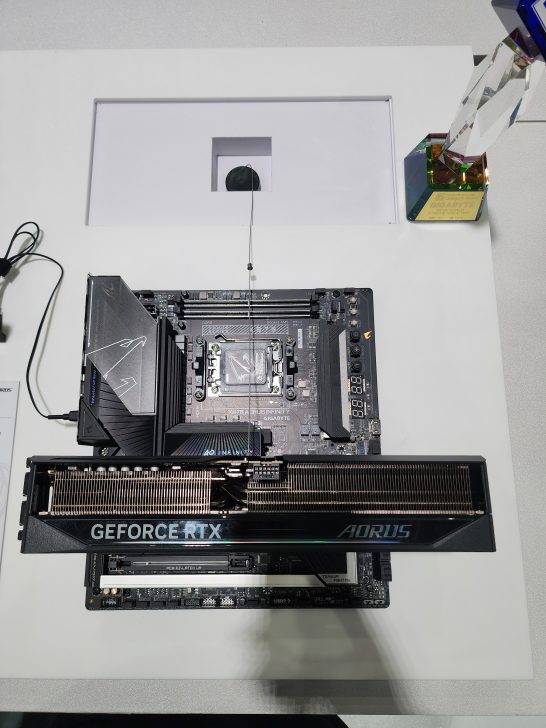 A Gigabyte AORUS motherboard with an AORUS GeForce RTX graphics card is displayed on a white surface next to a multicolored crystal trophy labeled 'GIGABYTE AORUS.'