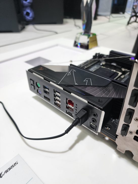 An AORUS branded motherboard with multiple USB ports and a connected cable is displayed on a table.