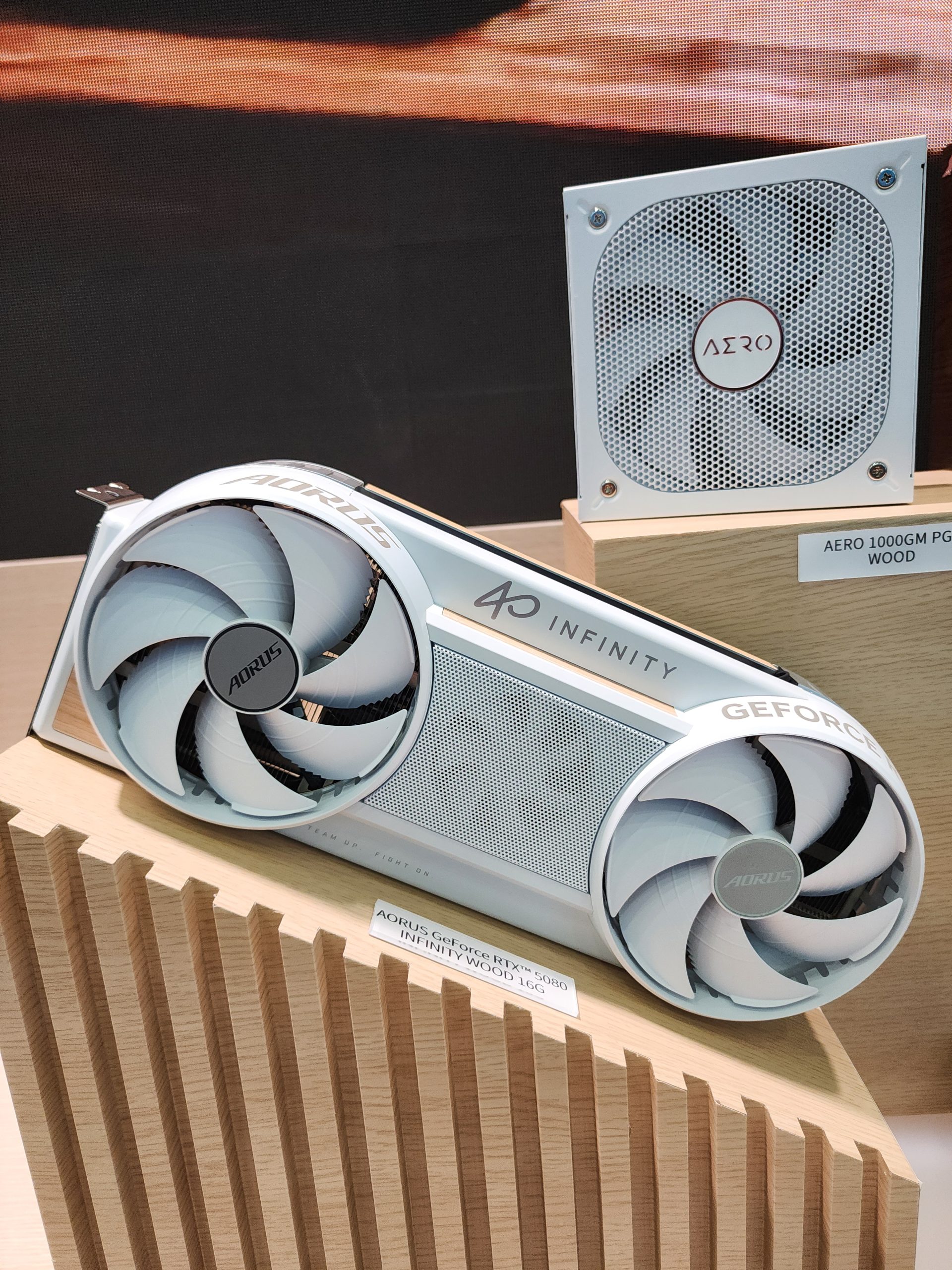 An AORUS GeForce RTX 5090 Infinity Wood 16G graphics card is displayed next to an AERO 1000GM PG5 WOOD power supply unit.