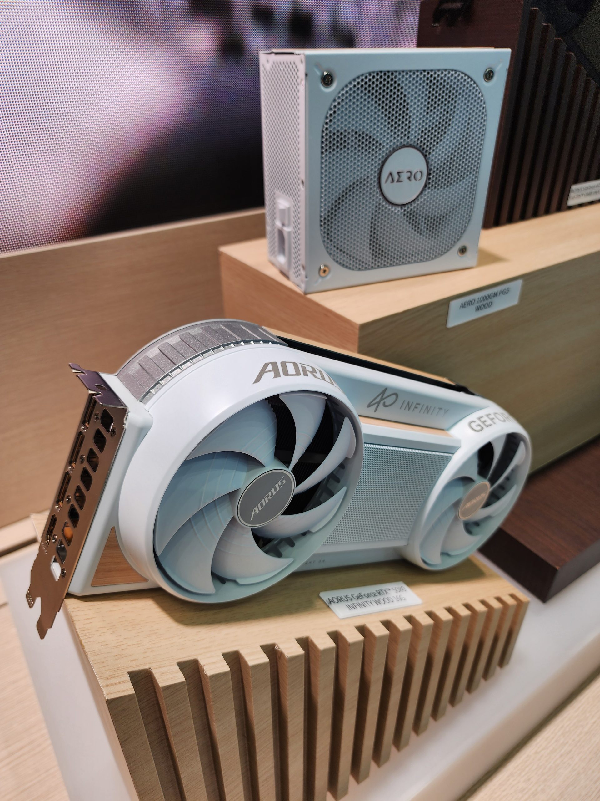 A white AORUS GeForce RTX 4090 INFINITY graphics card with dual fans is displayed next to an AERO 1000W power supply on a wooden stand.