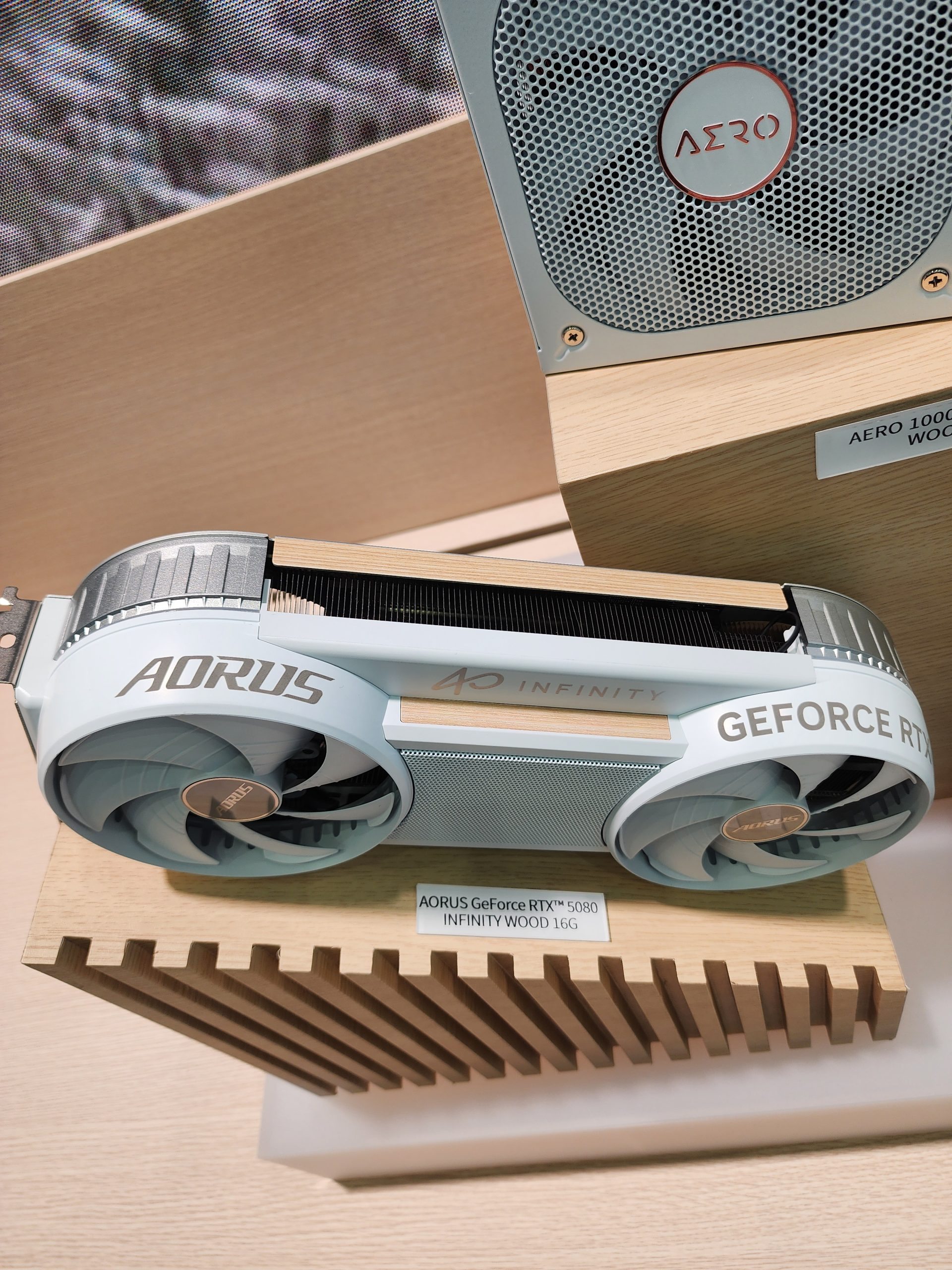 The AORUS GeForce RTX 5080 Infinity Wood 16G graphics card is displayed on a wooden stand next to an AERO 1000 series device.