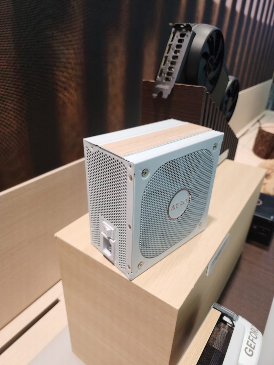 A white power supply unit labeled Aero is displayed on a wooden stand next to a partially visible graphics card with a black dual-fan design.