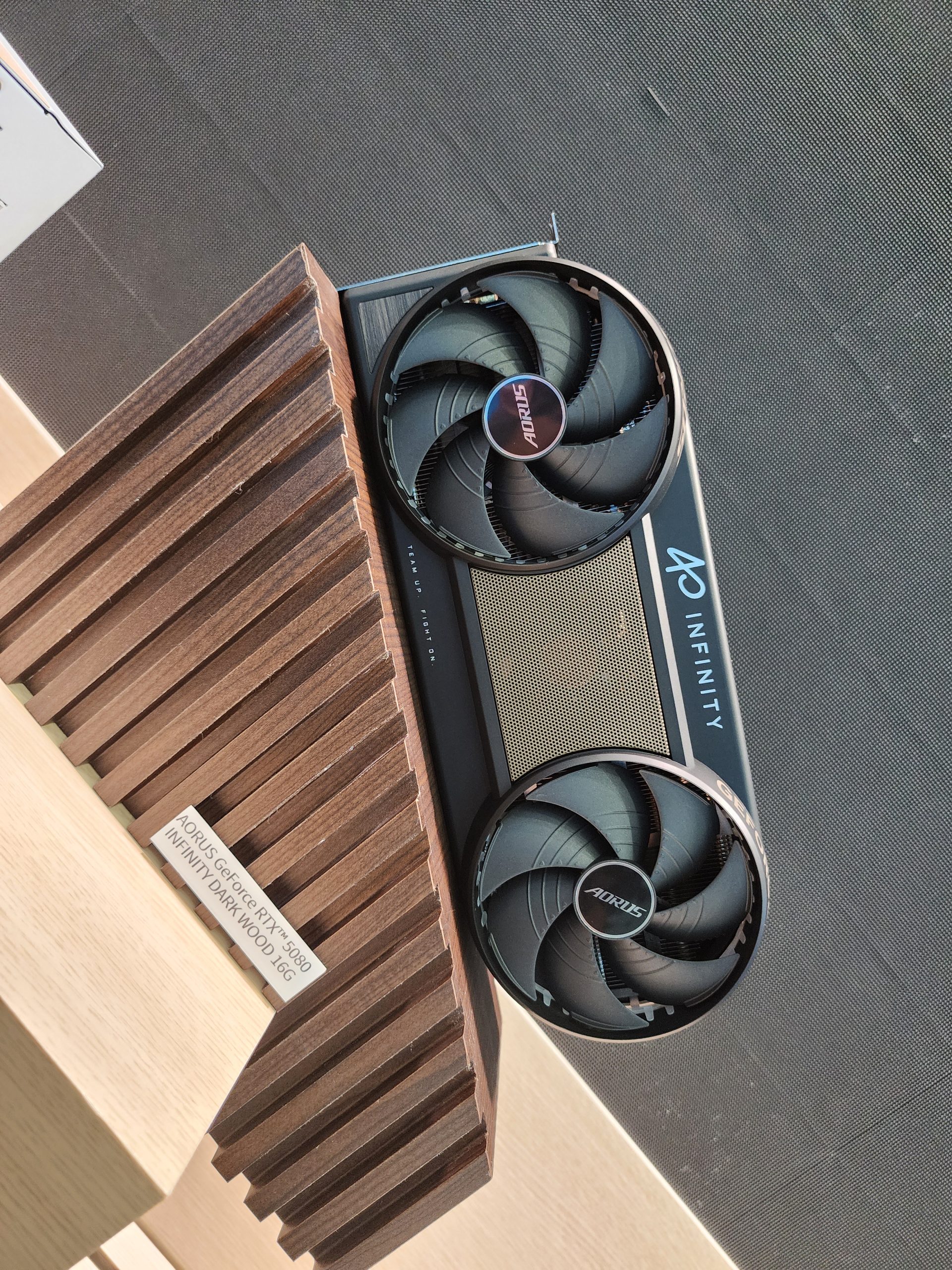 An AORUS GeForce RTX 5080 Infinity graphics card is displayed on a wooden stand labeled 'AORUS GeForce RTX 5080 Infinity Dark Wood 16G'.