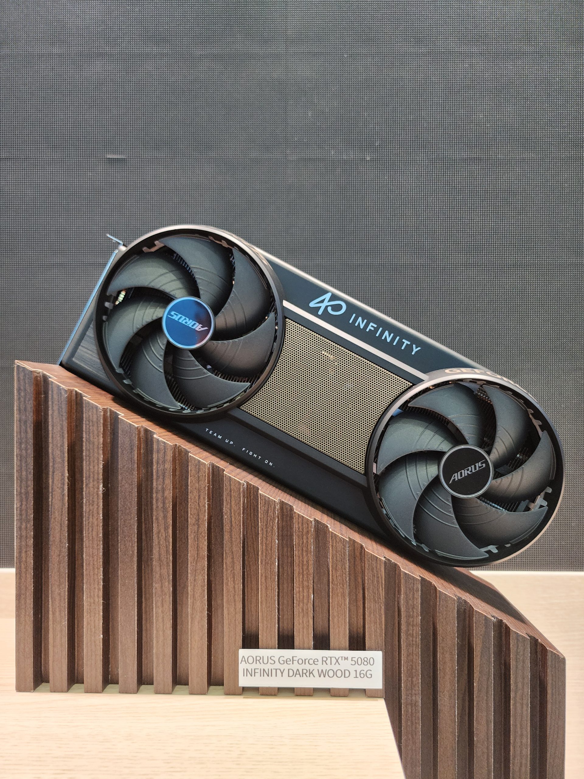 The AORUS GeForce RTX 5080 Infinity Dark Wood 16G graphics card is displayed on a wooden stand.