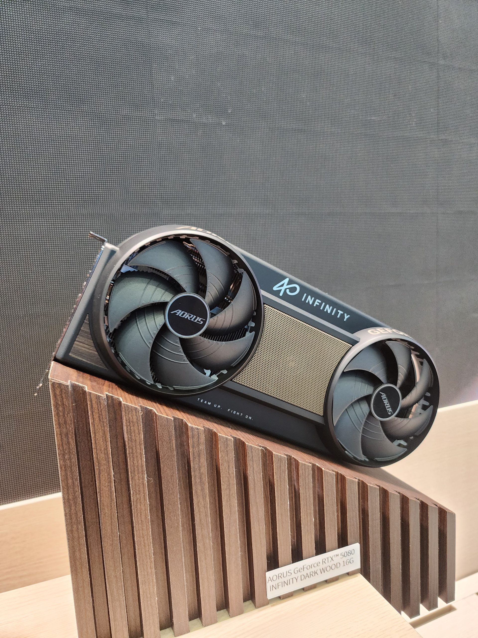 The AORUS GeForce RTX 5080 Infinity Dark Wood 16G graphics card is displayed on a wooden stand.