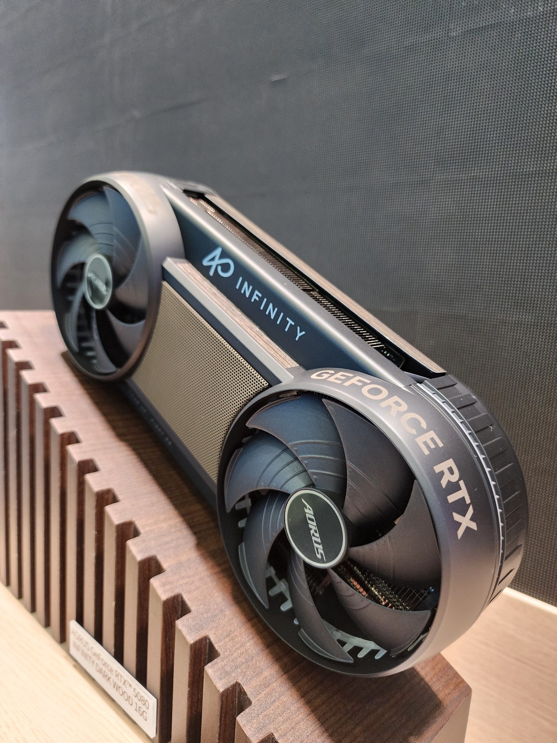 A GIGABYTE AORUS GeForce RTX 5090 graphics card is displayed on a wooden stand with visible branding 'INFINITY' and 'AORUS' on its fans.